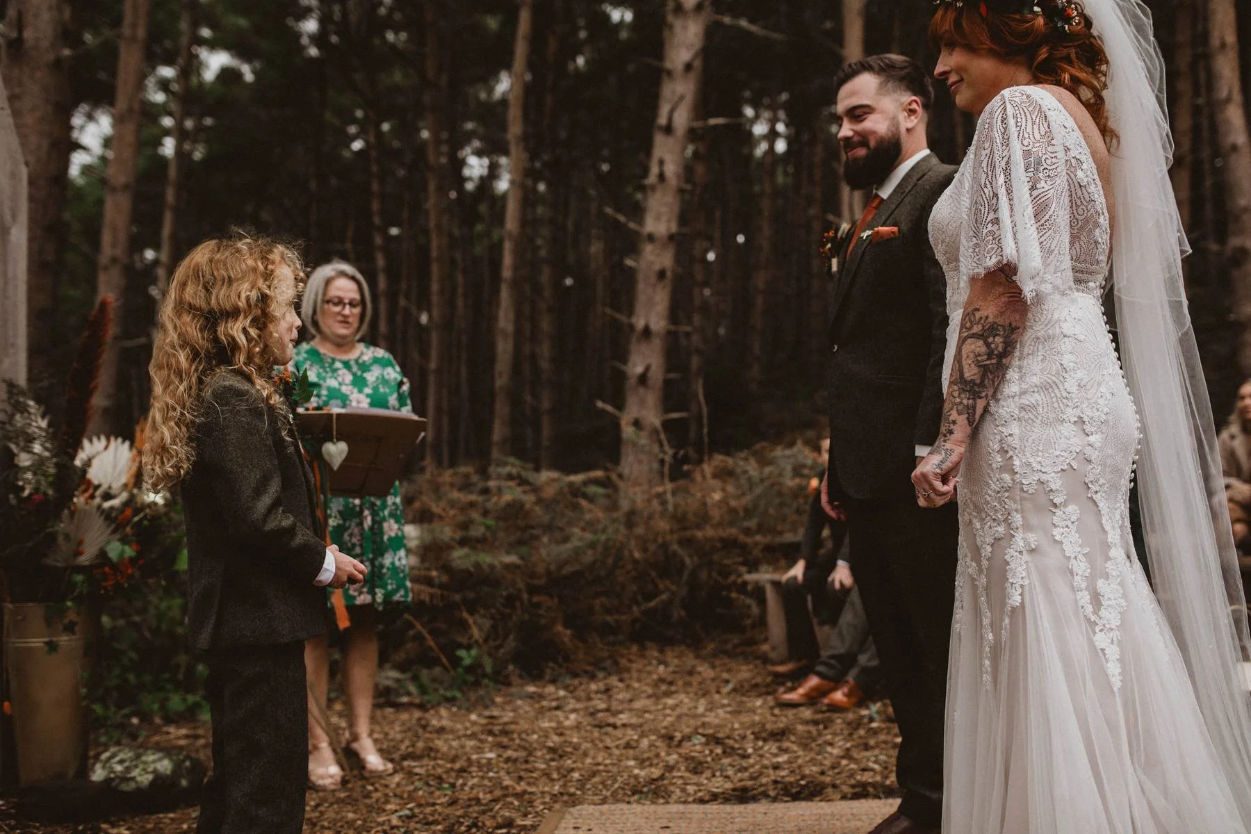 A wedding ceremony taking place in a forest with a bride and groom holding hands, facing a young child in a dark suit who appears to be presenting or speaking, with an officiant in a green dress standing behind them.