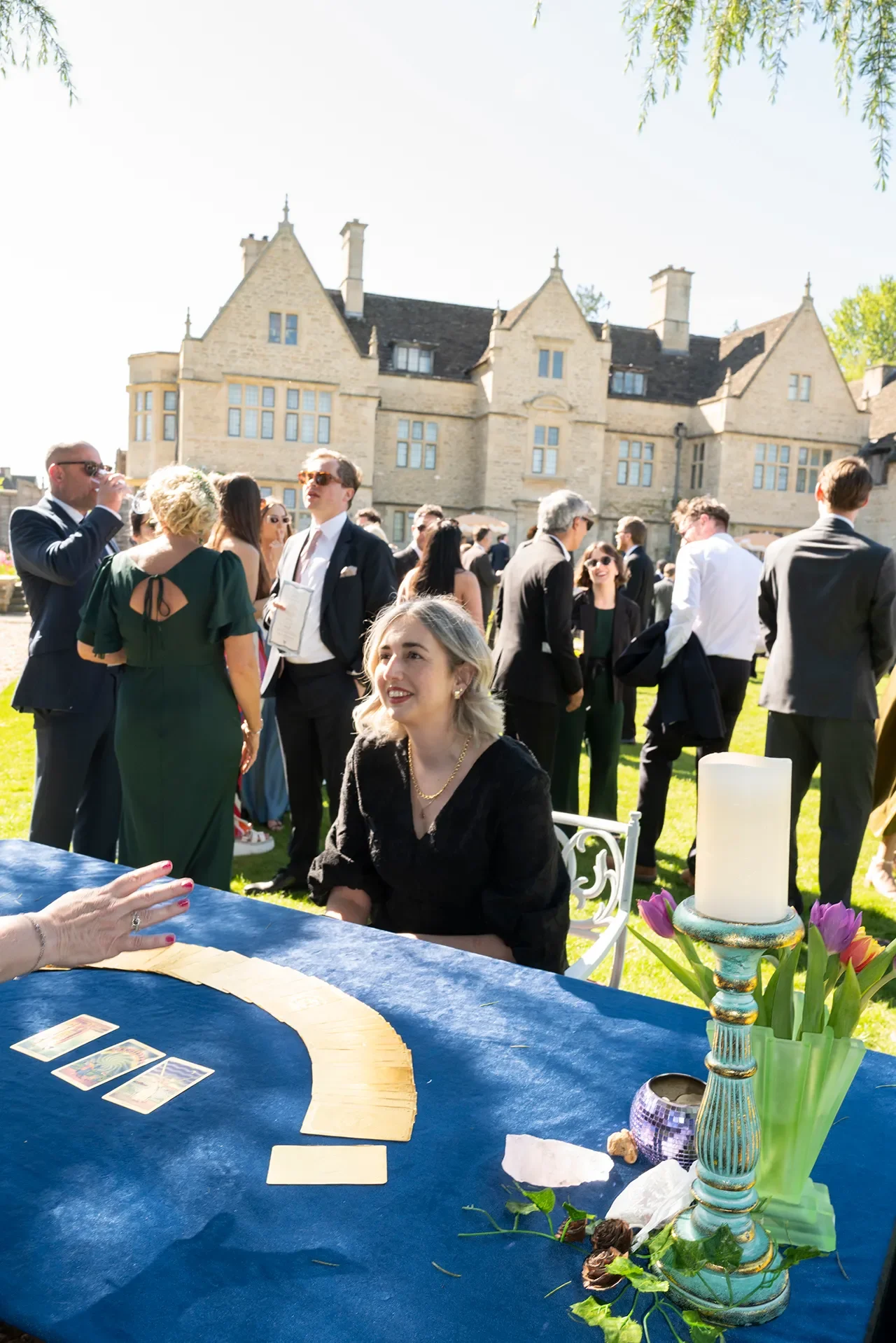 People gathered outdoors at a social event, with a large historic building in the background. A woman is sitting at a table with tarot cards, while others are mingling and talking, dressed in formal and semi-formal attire.