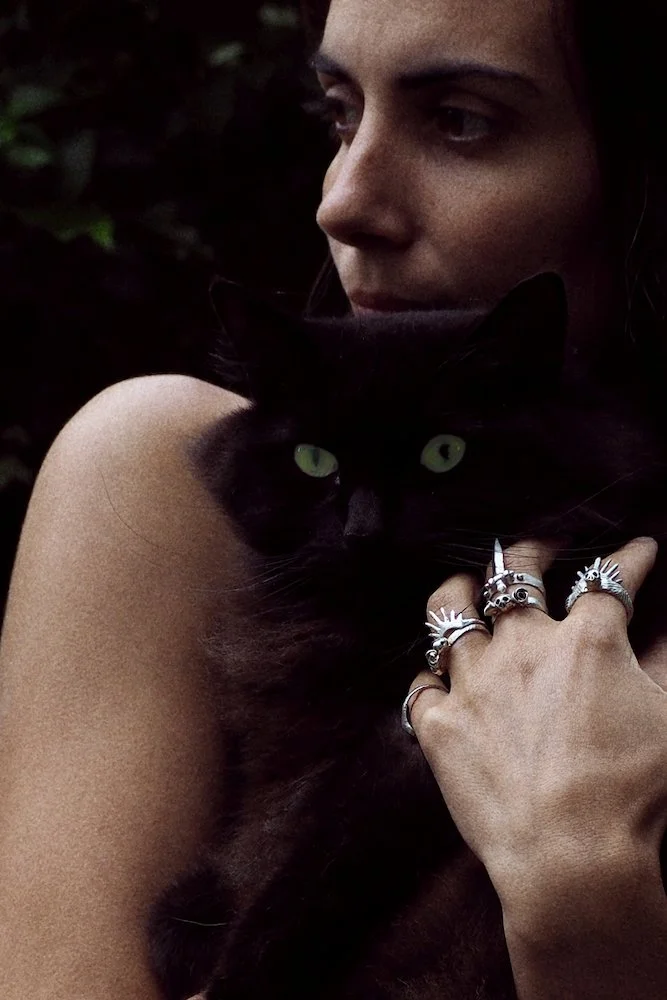 A woman with dark hair and multiple rings on her fingers holds a black cat with green eyes close to her face. The background is dark and blurred.