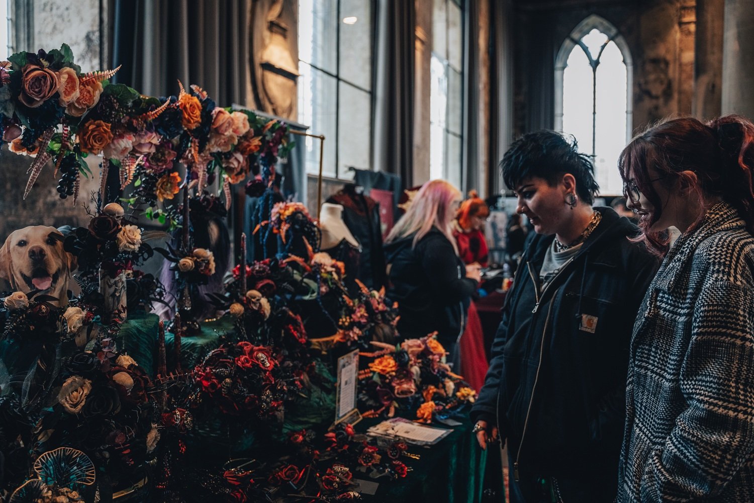 Two women shopping at a craft fair, looking at jewelry and accessories display. Mannequins and floral arrangements decorate the booth, with other vendors and shoppers in the background inside a high-ceiling, Gothic-style building with large arched windows.