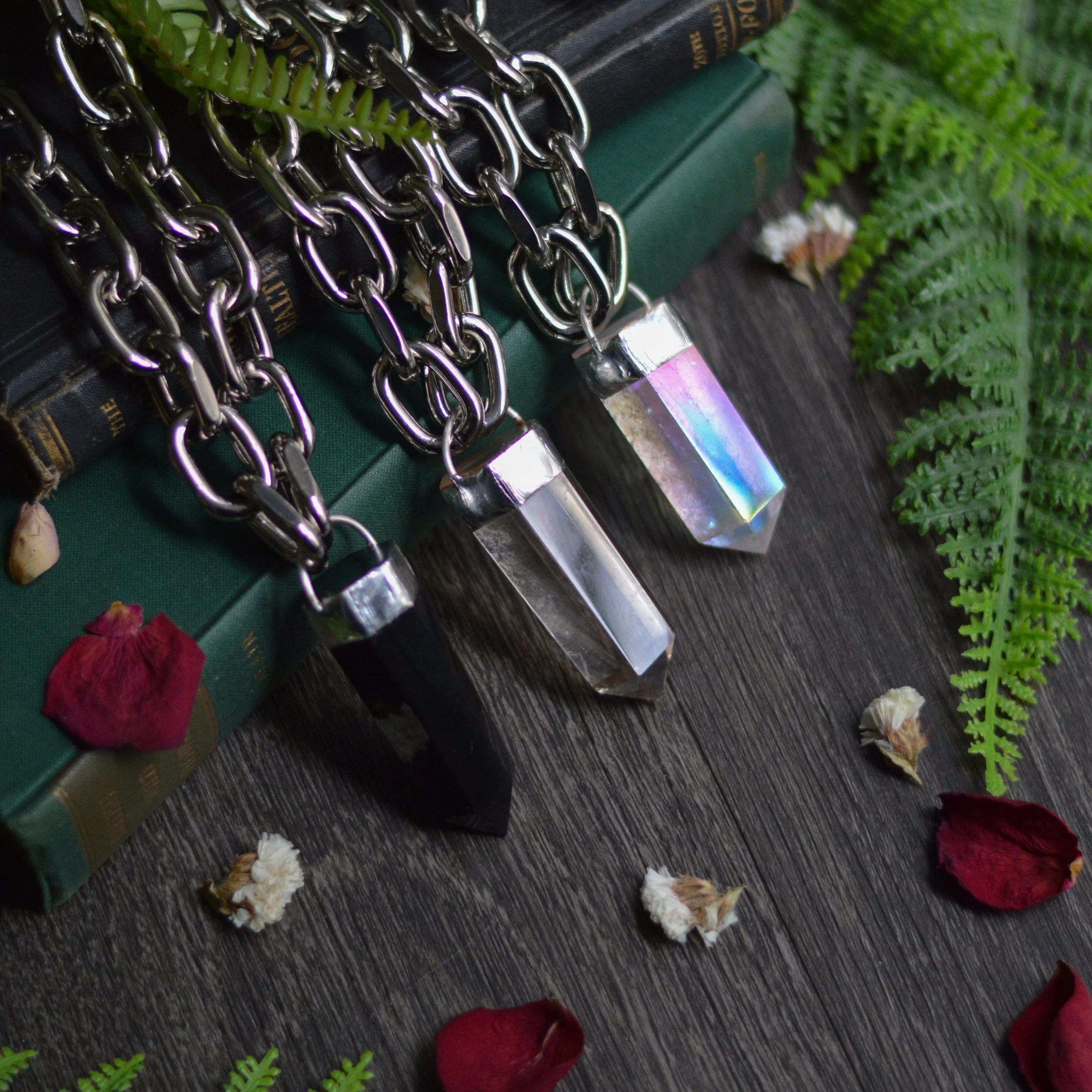 Three crystal pendants on silver chains laid on a dark wooden surface, with green fern leaves and dried flower petals surrounding them.