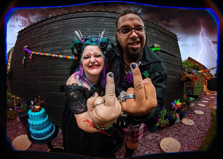 A woman with vibrant rainbow-colored hair, wearing gothic attire and accessories, and a man with glasses and a beard, dressed in punk style, pose closely with middle fingers raised in front of a dark wooden fence, with storm clouds and lightning in t