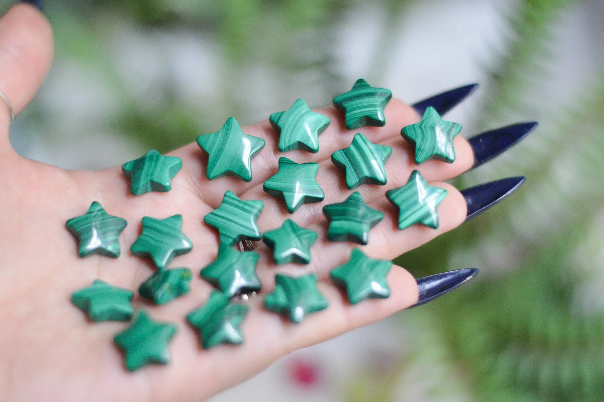 A hand with long, dark painted nails holds small green star-shaped objects made of marbled malachite stone.