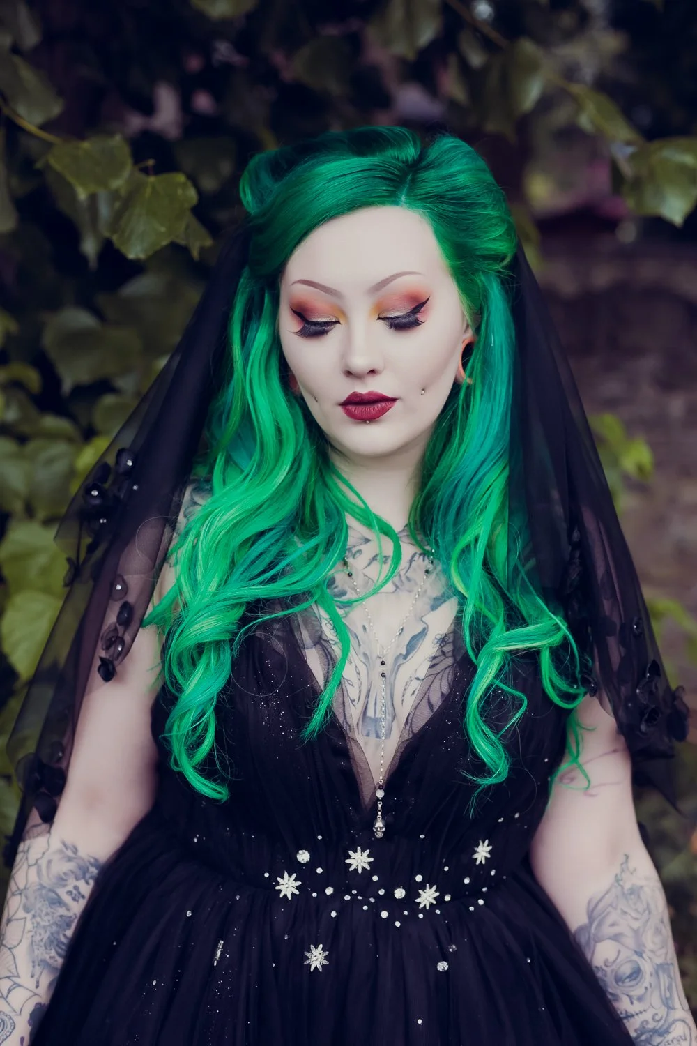 Woman with vibrant green hair, makeup, and tattoos, wearing a black dress with floral embroidery and a sheer overlay, standing outdoors against leafy background.