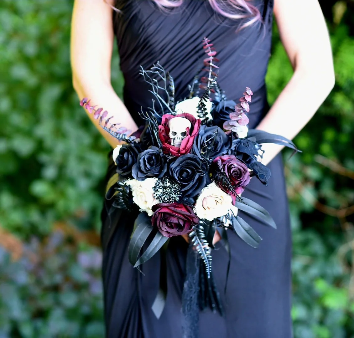 A person in a black dress holding a dark floral bouquet with black, purple, and white roses, black foliage, and small skull accents, set against a green outdoor background.