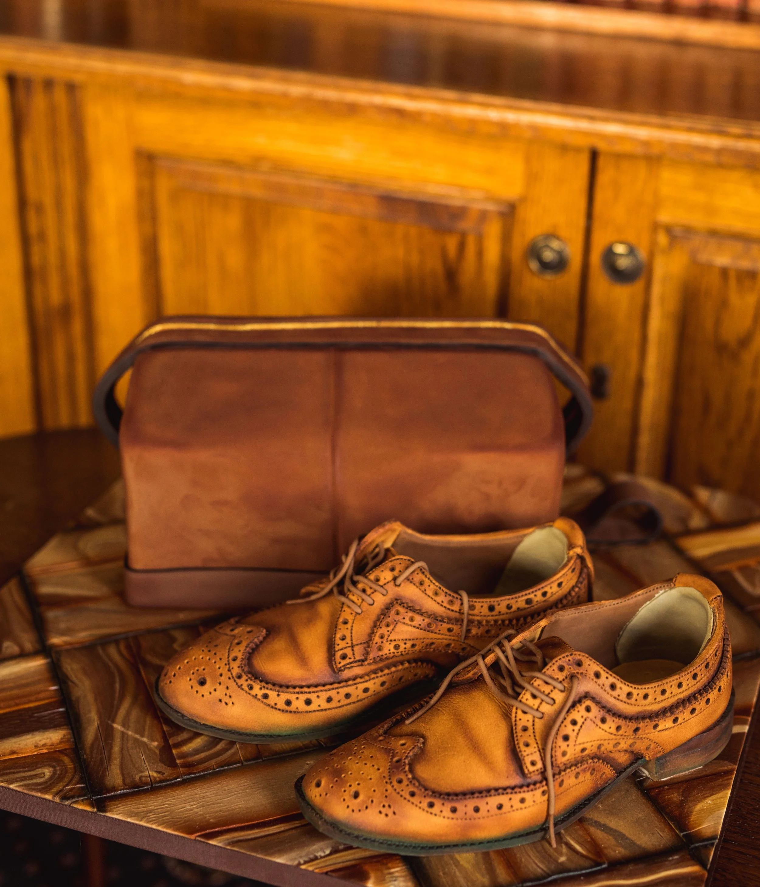 Brown leather brogue shoes, a pinkish-brown bag, and a brown leather wallet placed on a wooden table with a wooden cabinet in the background.