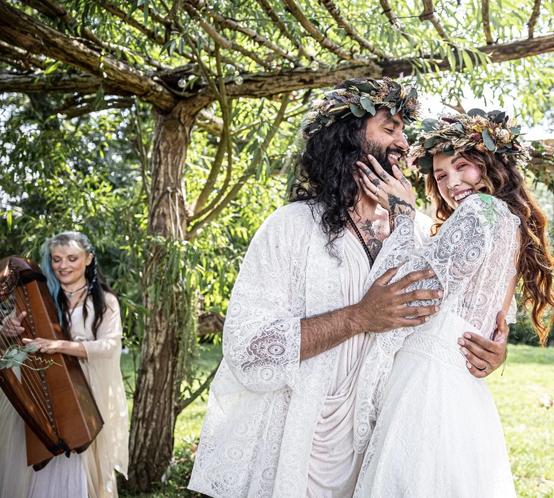 A couple is dressed in white, smiling and embracing under a tree, with a woman playing the harp in the background, during a wedding or celebration in an outdoor garden setting.