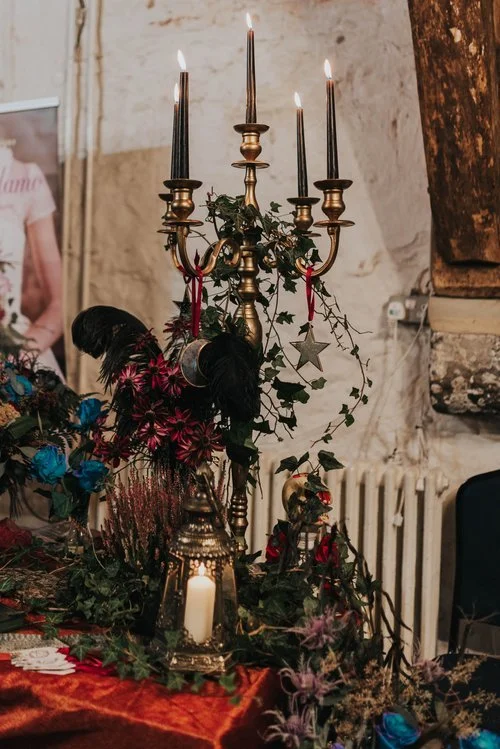 A vintage brass candelabrum with five lit candles, decorated with green vines, red ribbons, and star-shaped ornaments. It is surrounded by colorful flowers, including pink and blue blossoms, and placed on a table covered with a red cloth. The background features a rustic wall and a white radiator.
