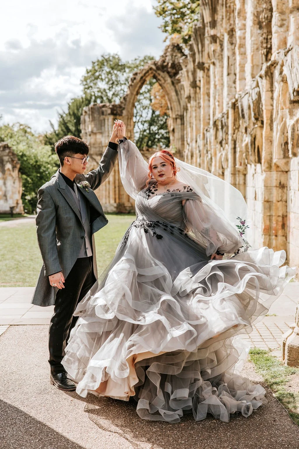 A couple in wedding attire dancing outdoors near ancient ruins with stone archways and green trees.