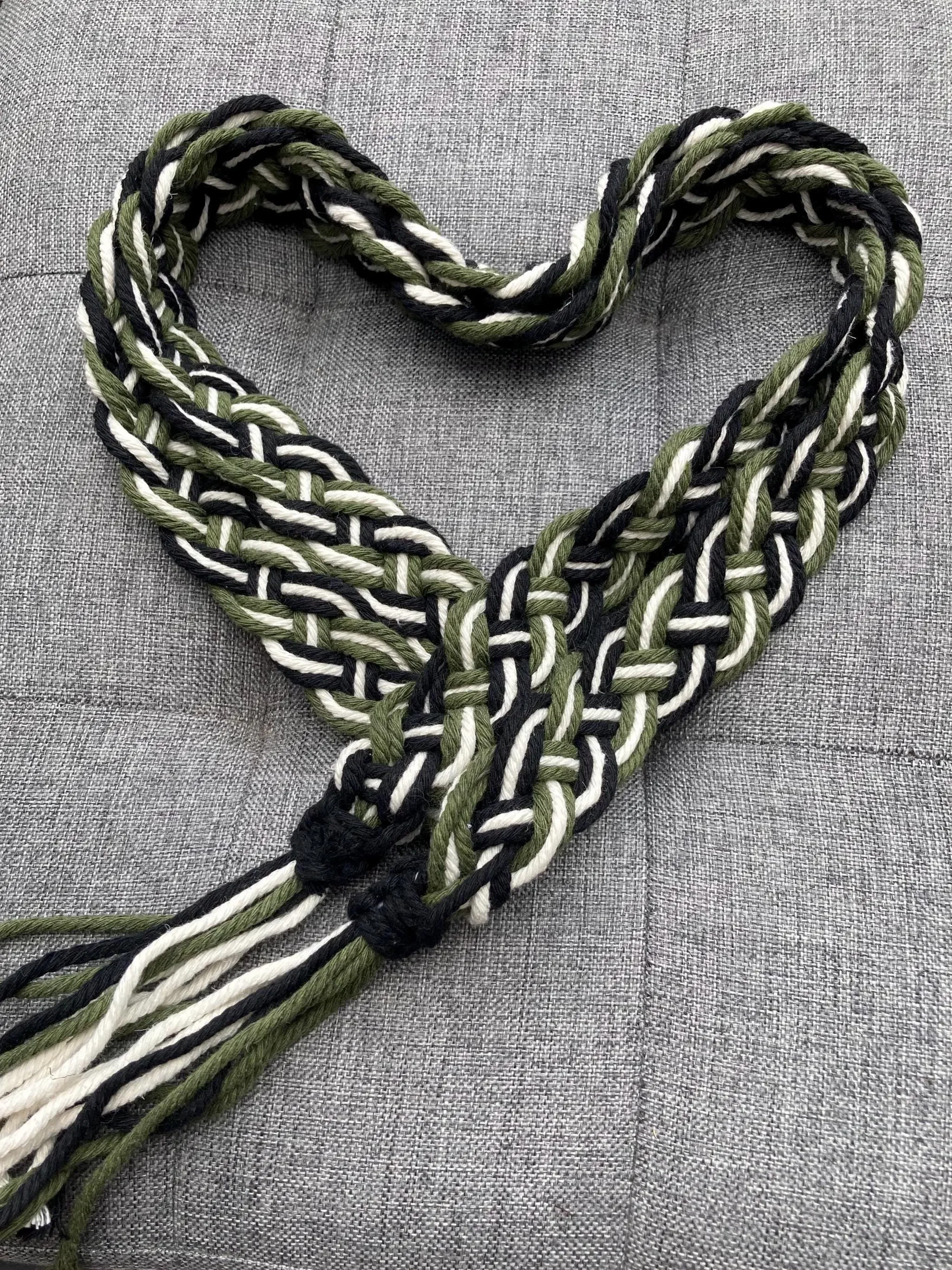 A heart-shaped arrangement of woven green, black, and white yarns on a gray fabric background.