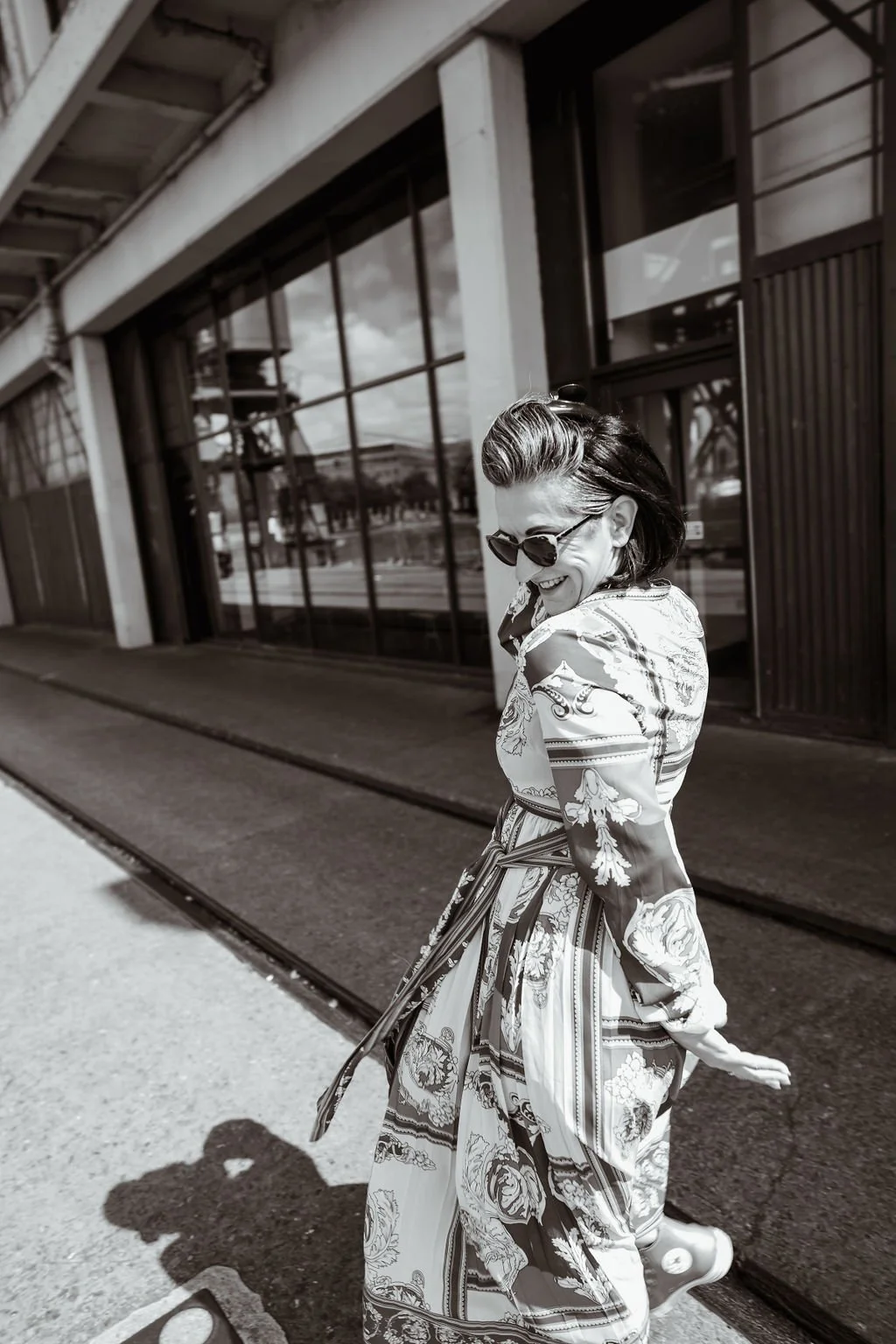 A woman with short dark hair wearing sunglasses and a patterned dress smiling as she walks along a sidewalk in front of a modern building with large glass windows.