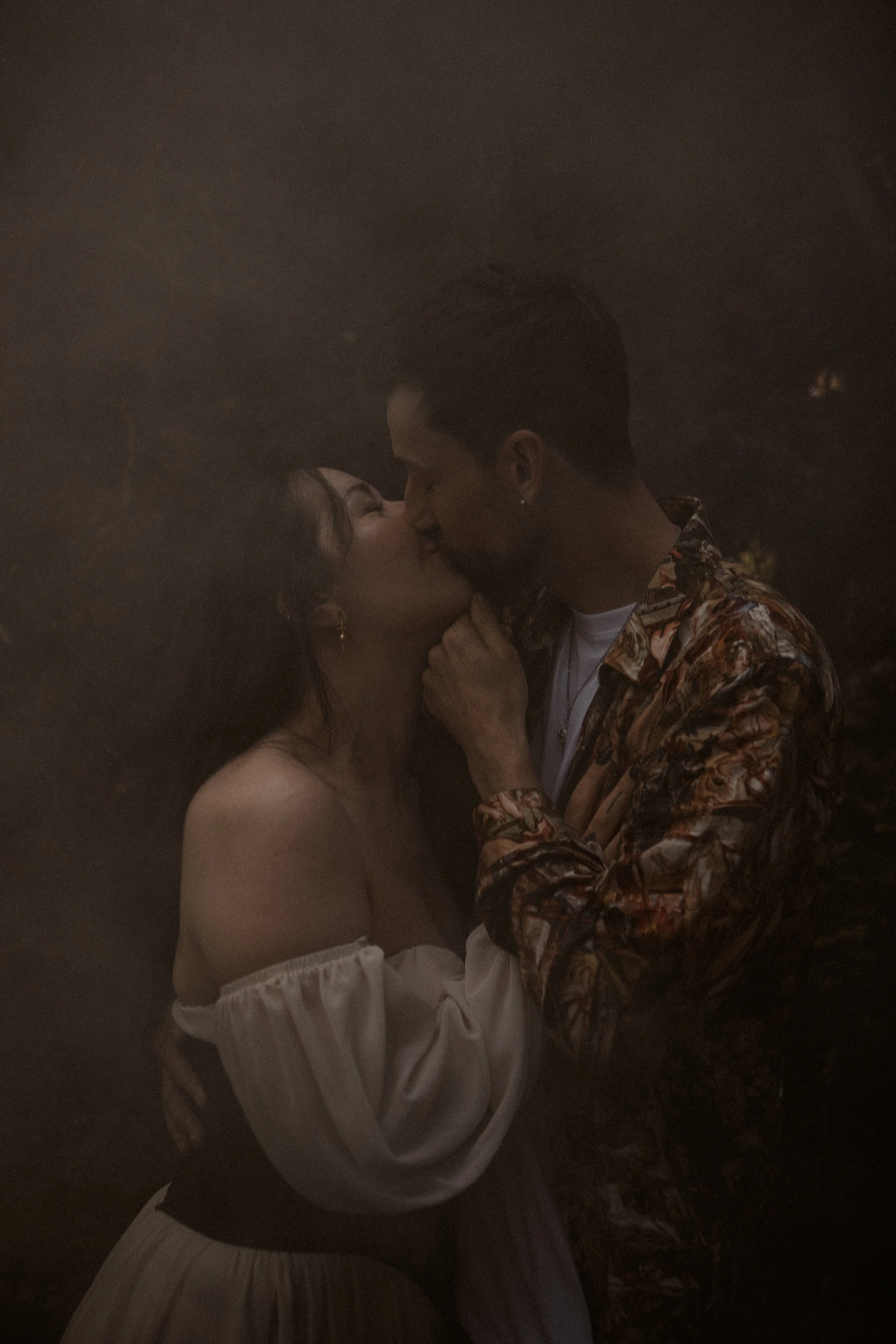 A couple sharing a kiss, with the woman wearing an off-shoulder dress and the man wearing a patterned shirt.