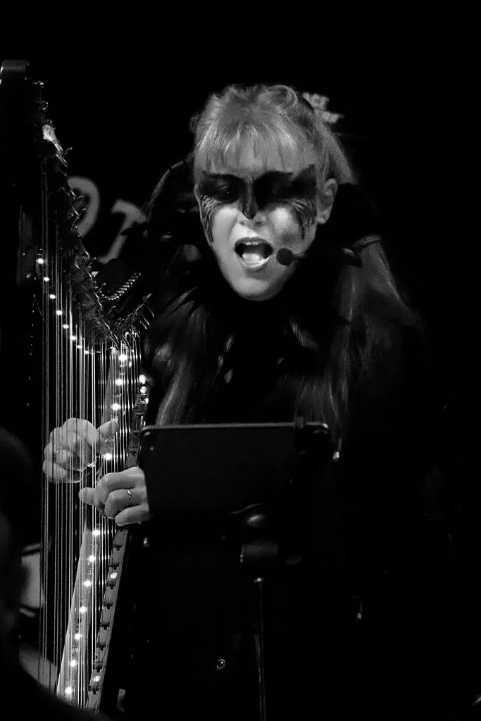 A black and white photo of a woman singing into a microphone while playing a harp decorated with string lights.