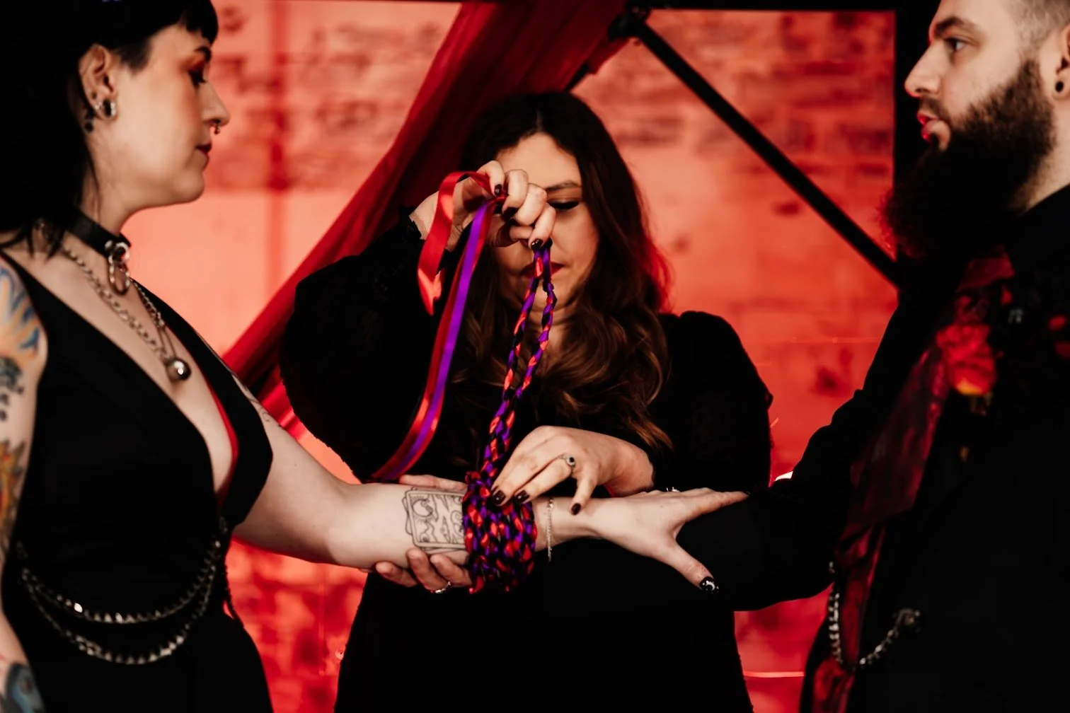 A couple is partaking in a wedding ceremony, with an officiant tying a red and purple ribbon around their wrists, symbolizing their union, in a setting with a warm, red brick background.