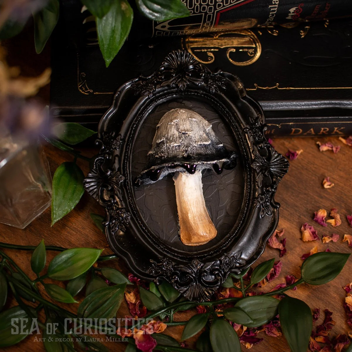 A dark decorative oval frame contains a dried mushroom with a dark cap and light stem. The frame and mushroom are on a wooden surface, surrounded by green leaves and scattered dried pink flower petals. A book with dark cover and gold lettering is in 