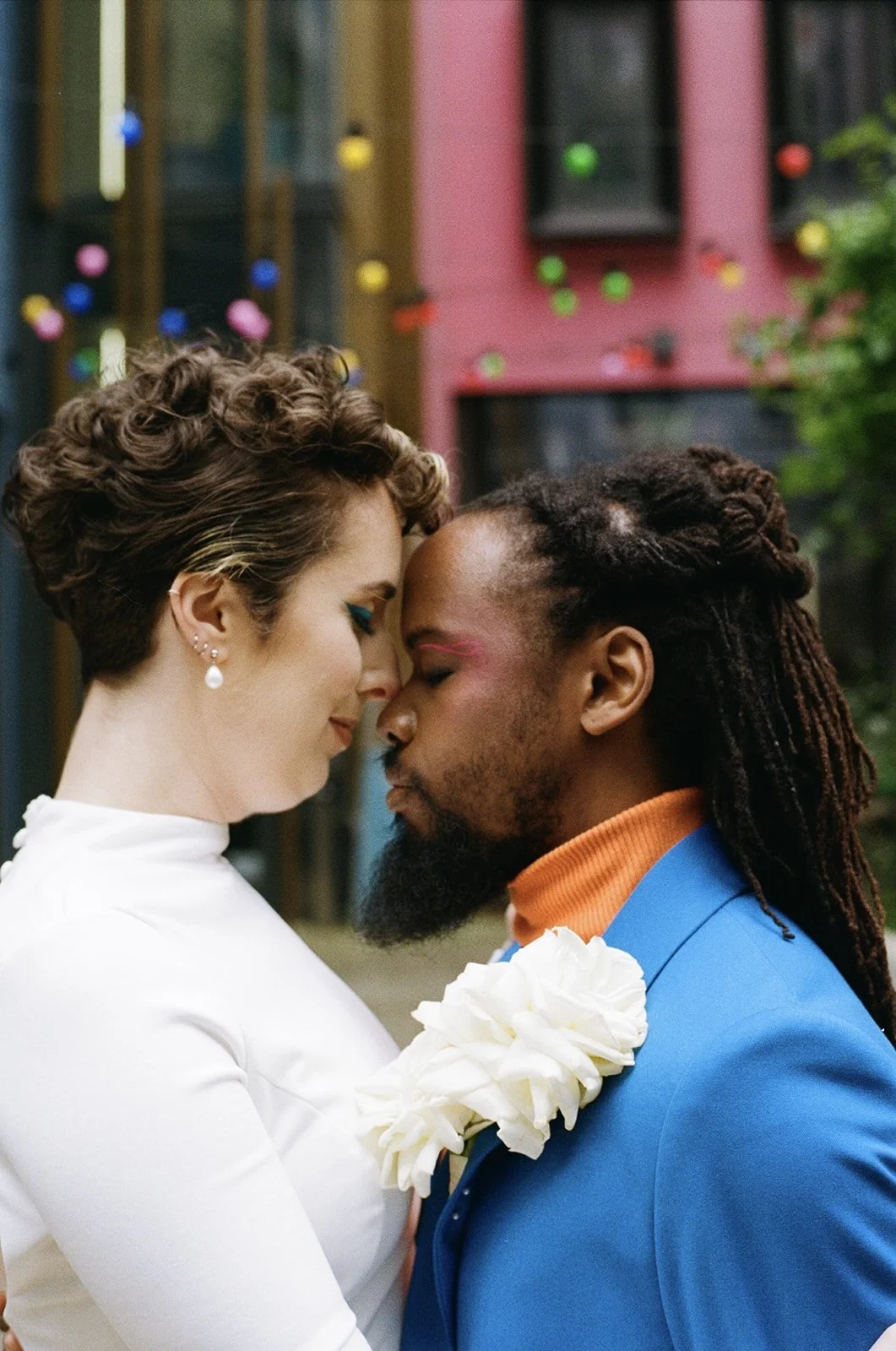 A close-up of a couple with foreheads touching, eyes closed, outdoors in front of colorful buildings and string lights, expressing intimacy.