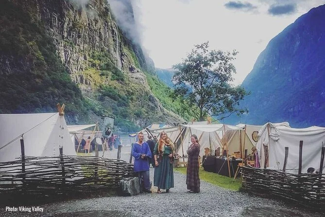 Viking village built from willow fences and white canvas tents. £ people in traditional viking dress face teh camera. Dramatic cliffs surround them