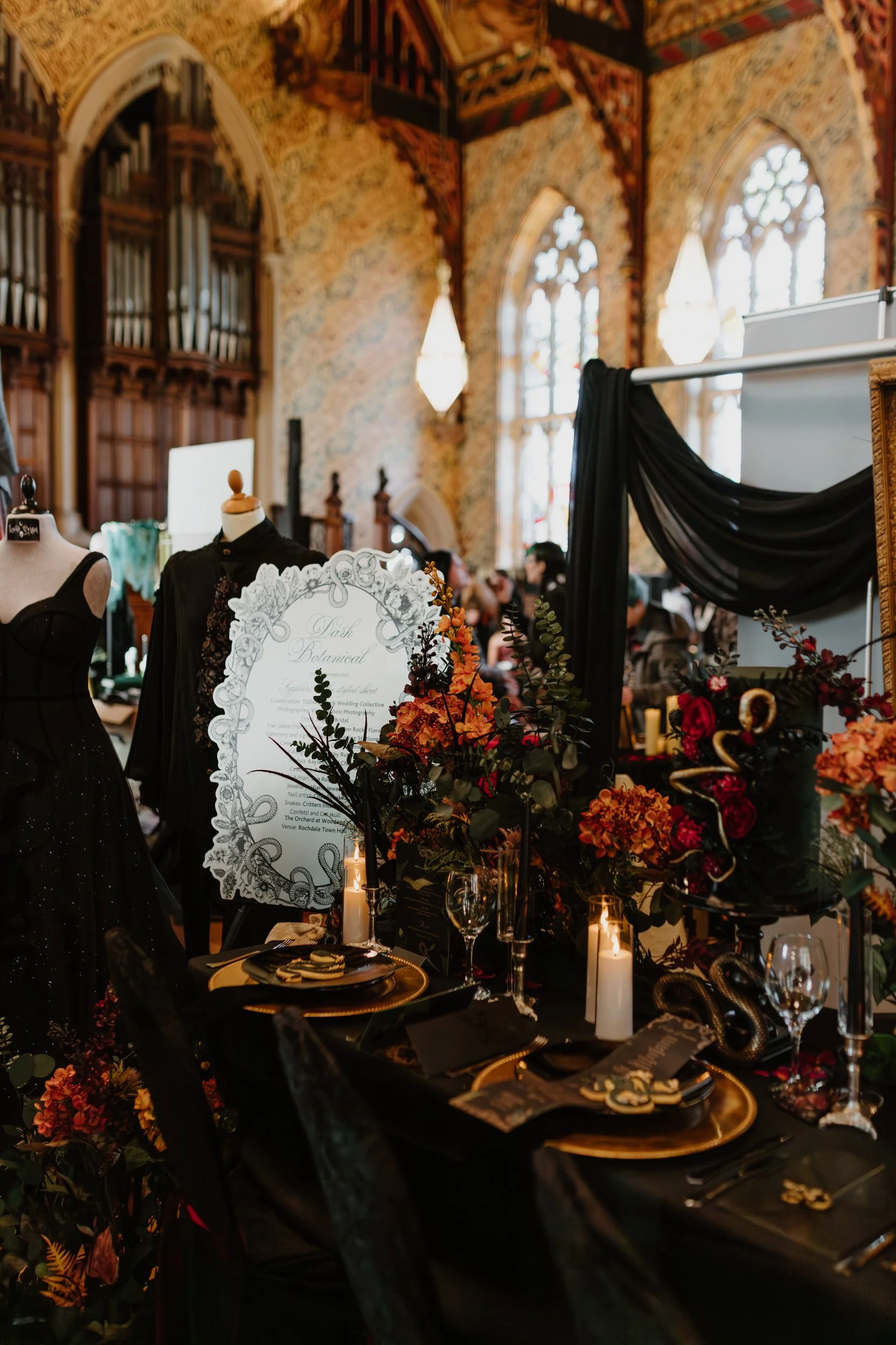 A richly decorated event space with gothic architecture, floral arrangements, candles, and black-themed table settings, featuring mannequins dressed in black clothing.