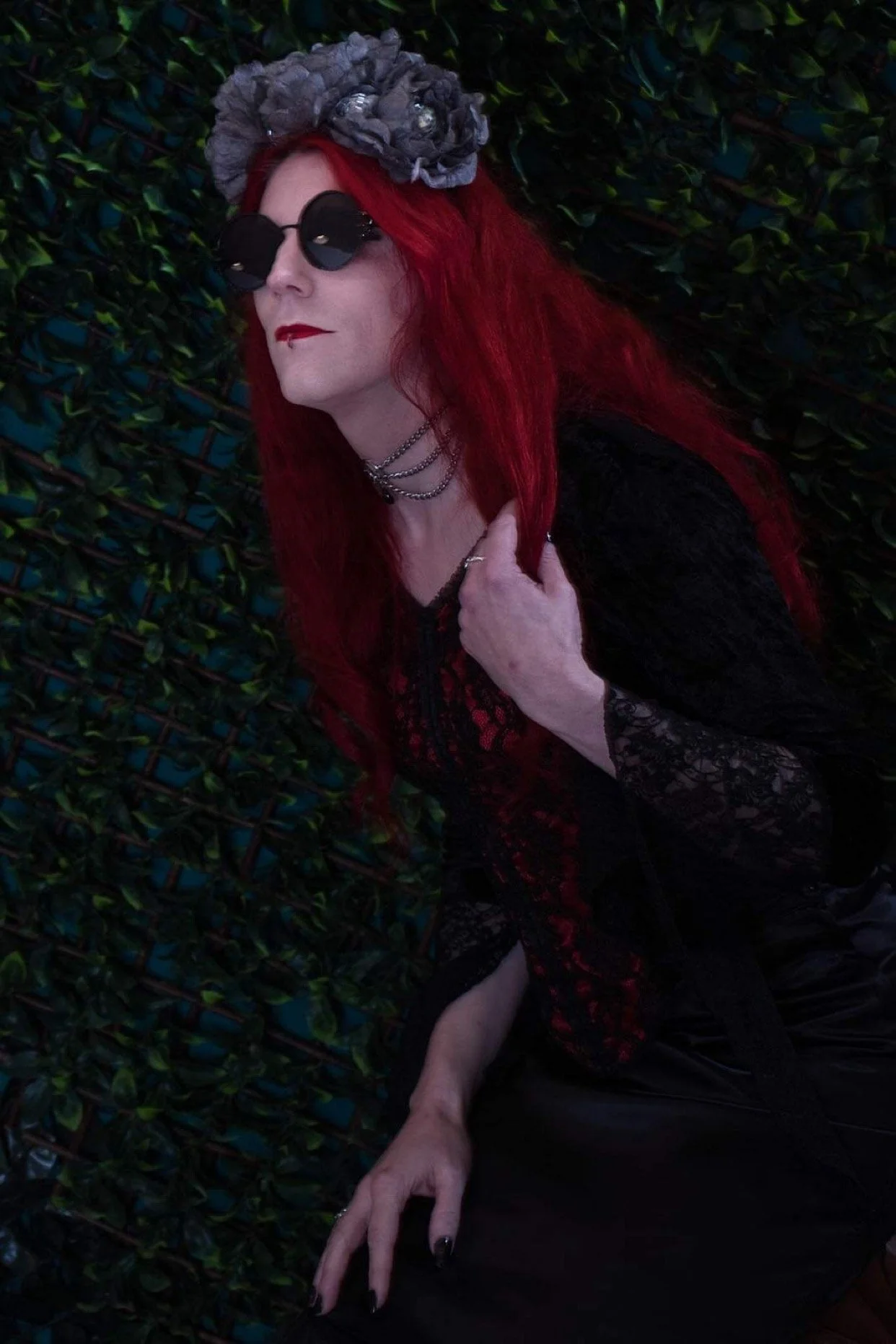Person with long red hair wearing black sunglasses and a gray floral headpiece, dressed in black lace clothing, standing against a green leafy background.