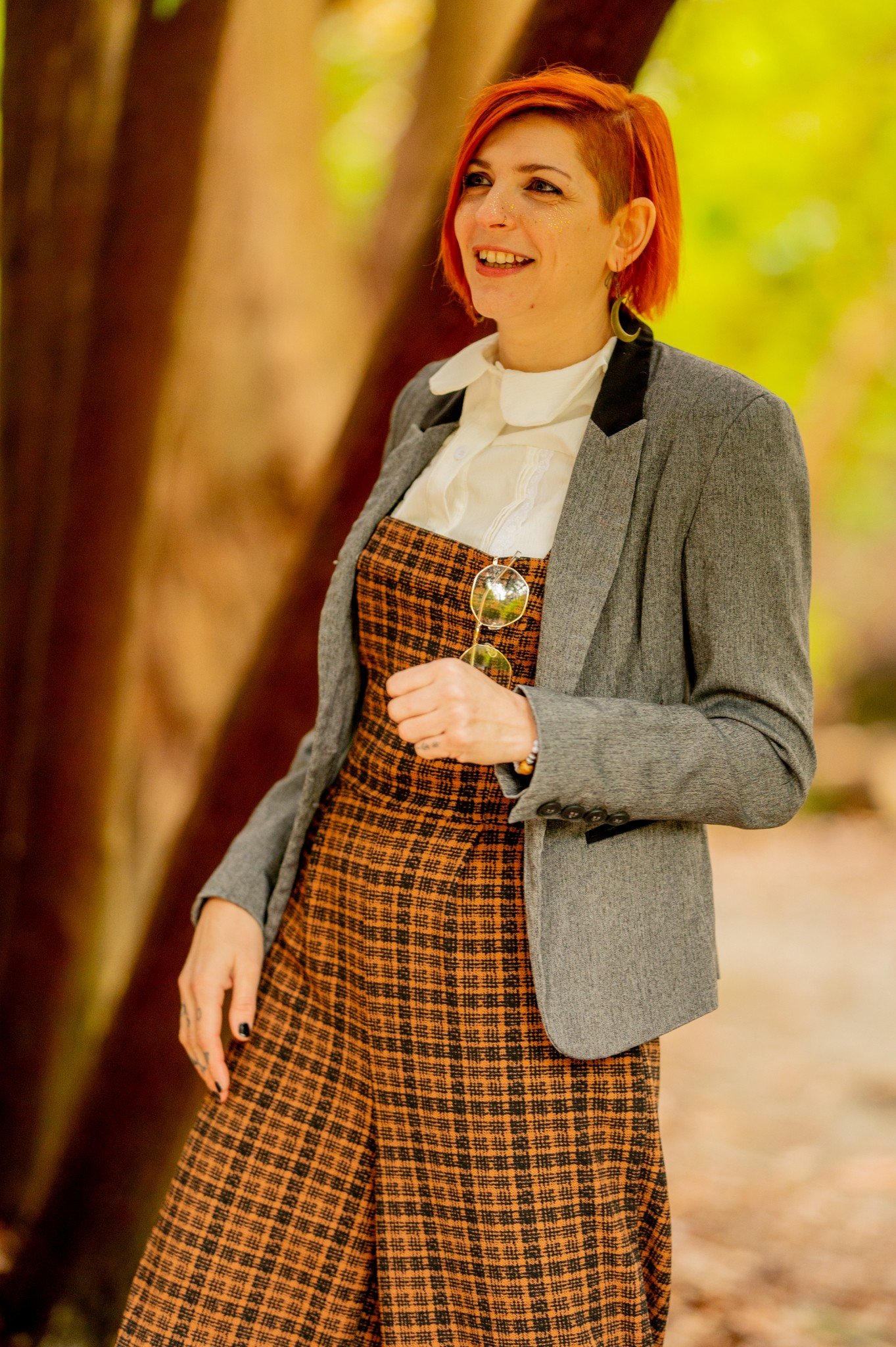 A woman with short orange hair, wearing a gray blazer over a white blouse, and a patterned brown and black skirt, holding sunglasses, standing outdoors among trees with blurred green and brown background.