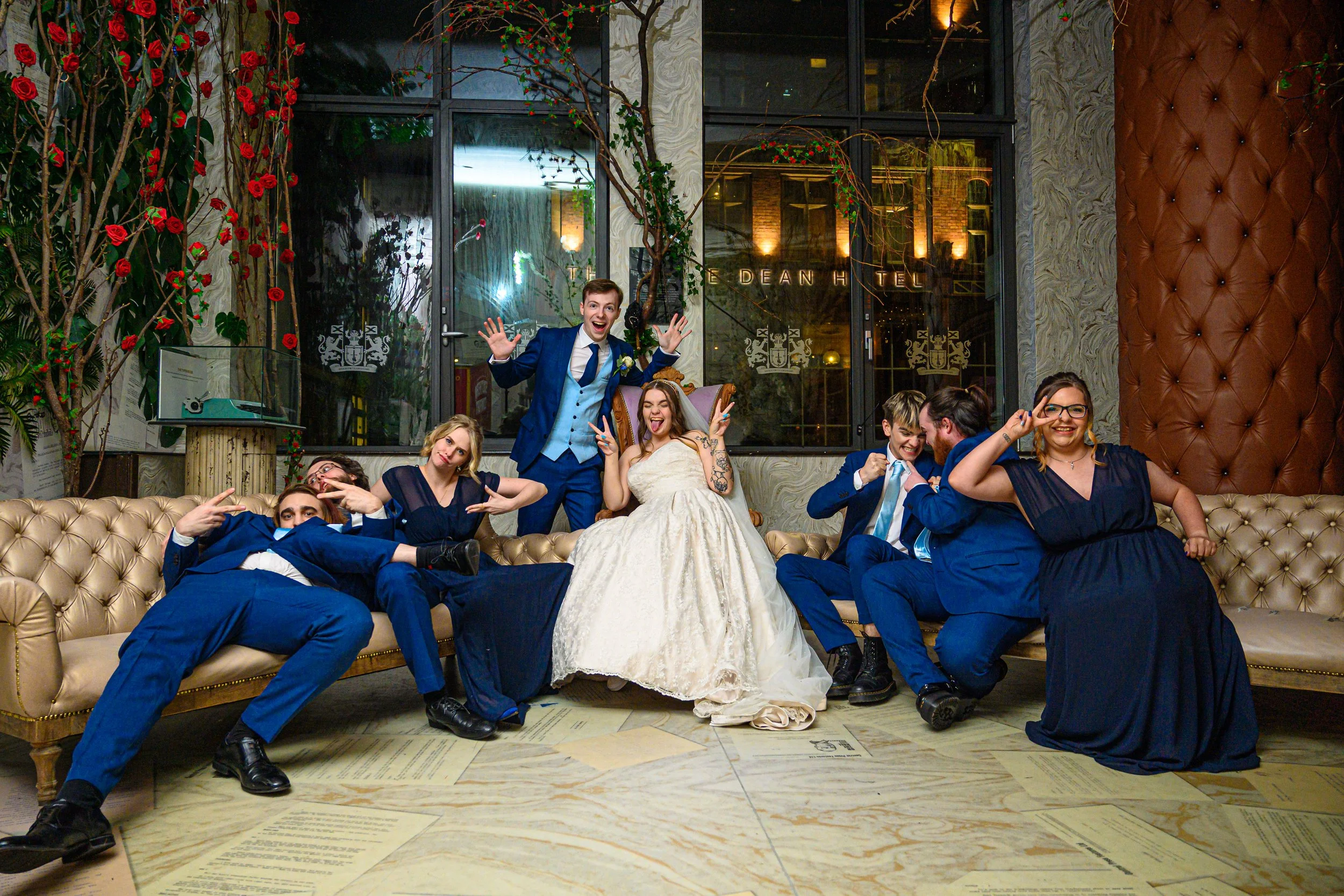 Group of wedding guests in formal attire posing playfully on a vintage sofa indoors, with a bride and groom at the center, making funny gestures and expressions.