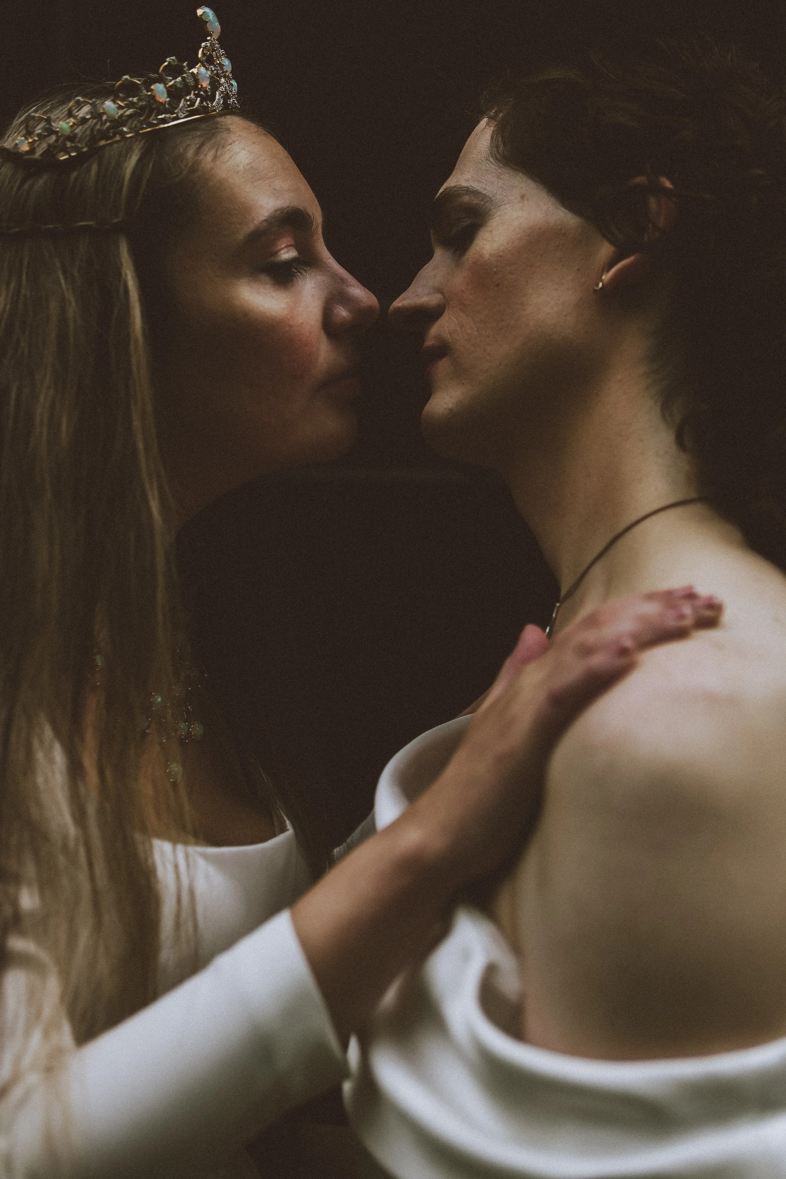 A close-up photo of two women touching foreheads, one wearing a crown and formal attire, the other with tattoos and dark makeup, in an intimate embrace.