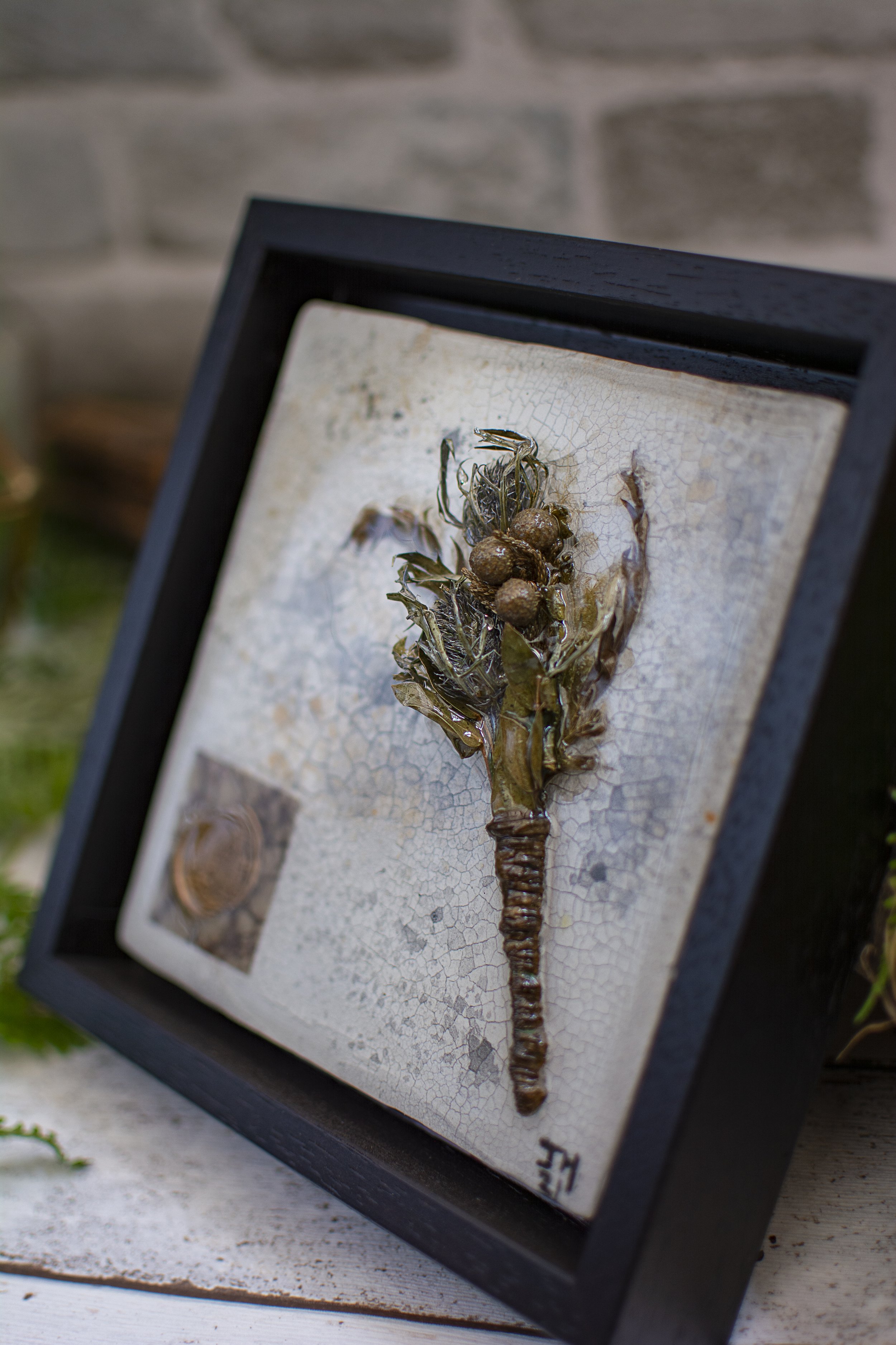 Framed art piece of a tree made from metallic materials, mounted on a cracked white background, leaning against a brick wall.