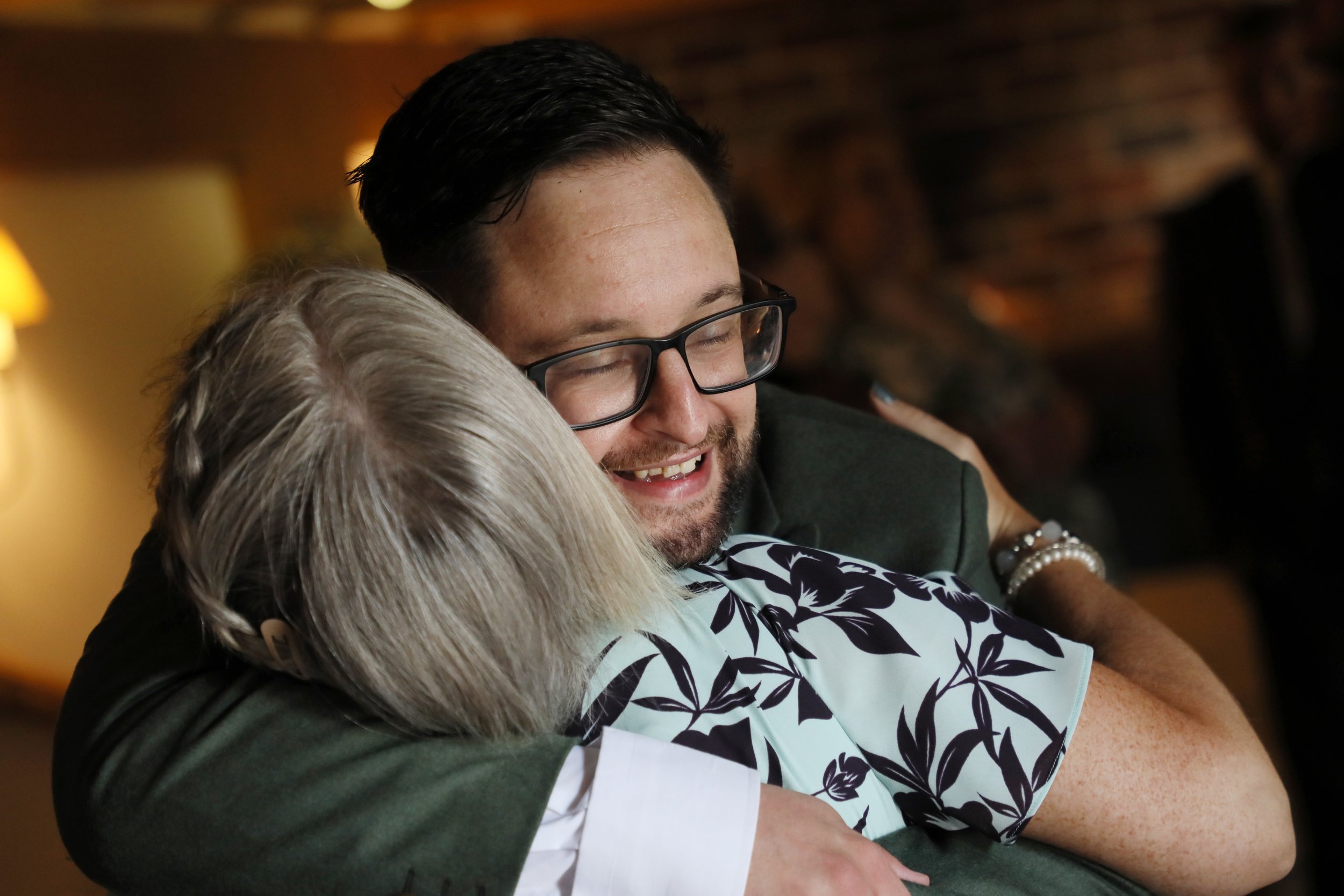 A man with glasses hugging an elderly woman with gray hair, both smiling warmly.