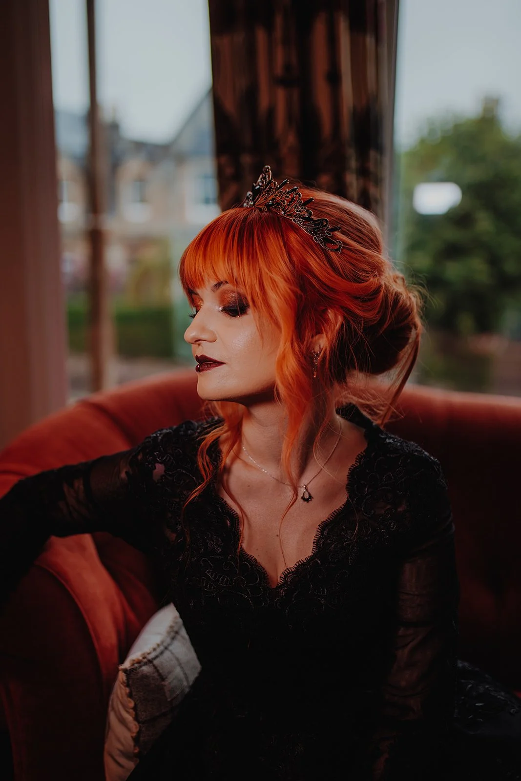 A woman with fiery red hair, wearing a black lace dress and a decorative black tiara, sitting on a vintage-style sofa, with a window and trees in the background.