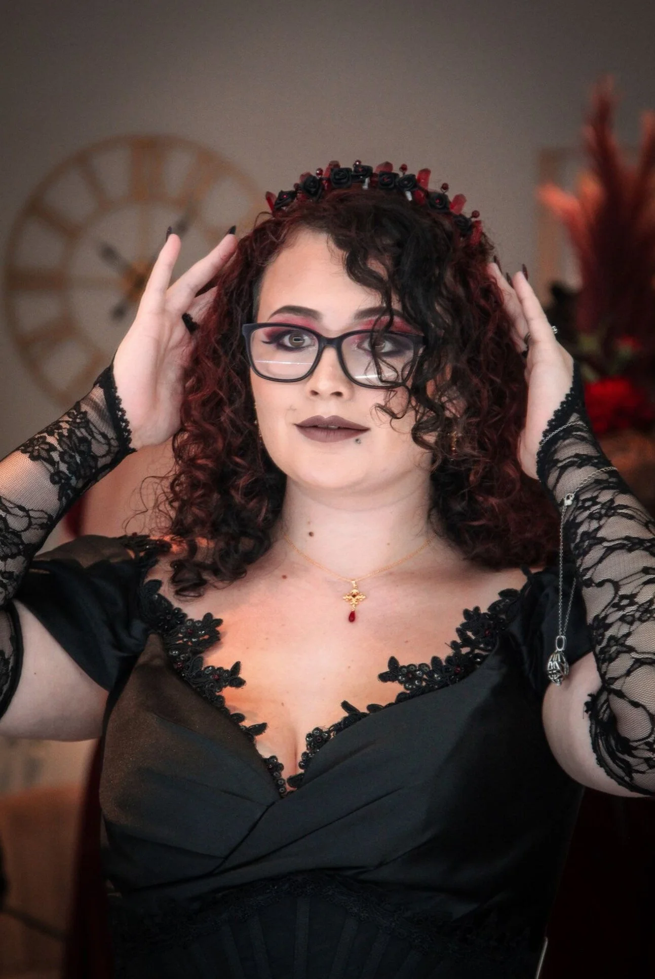A woman with curly dark hair wearing glasses, black lace top, and a red and black floral headband, adjusting her glasses indoors.