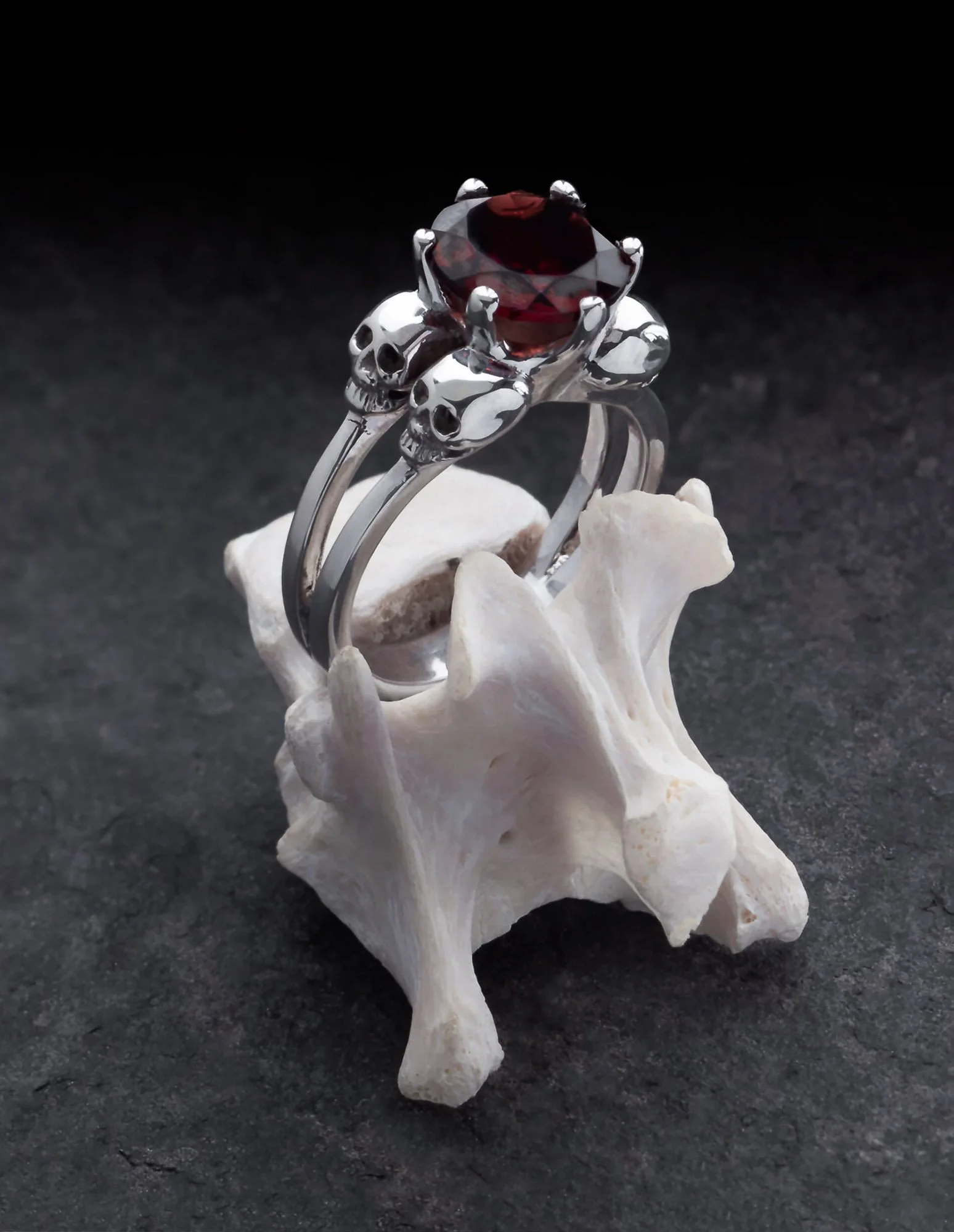 A silver ring with skull designs and a large red gemstone, displayed on a white skull-shaped object against a black background.