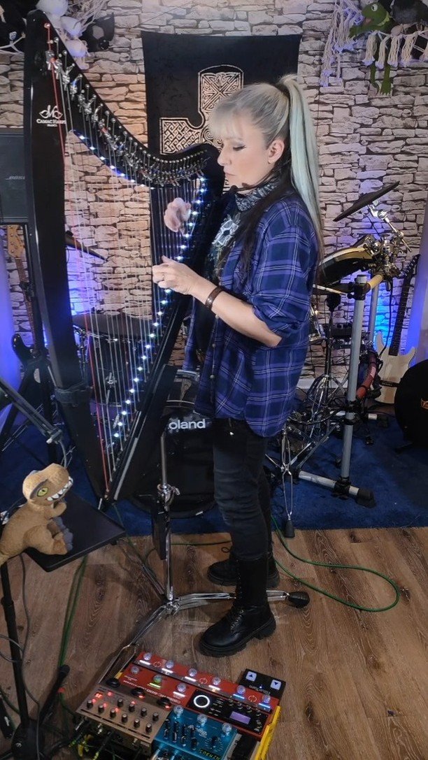 A woman with long blonde hair playing a harp decorated with string lights in a music studio with brick walls, other musical instruments, and recording equipment.