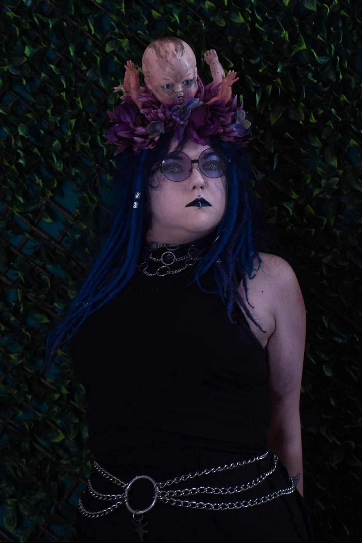 Person with long blue hair, black makeup, and sunglasses, wearing chained accessories, lying against a dark green leafy background, with a unique headpiece featuring a doll's head, purple flowers, and fabric.