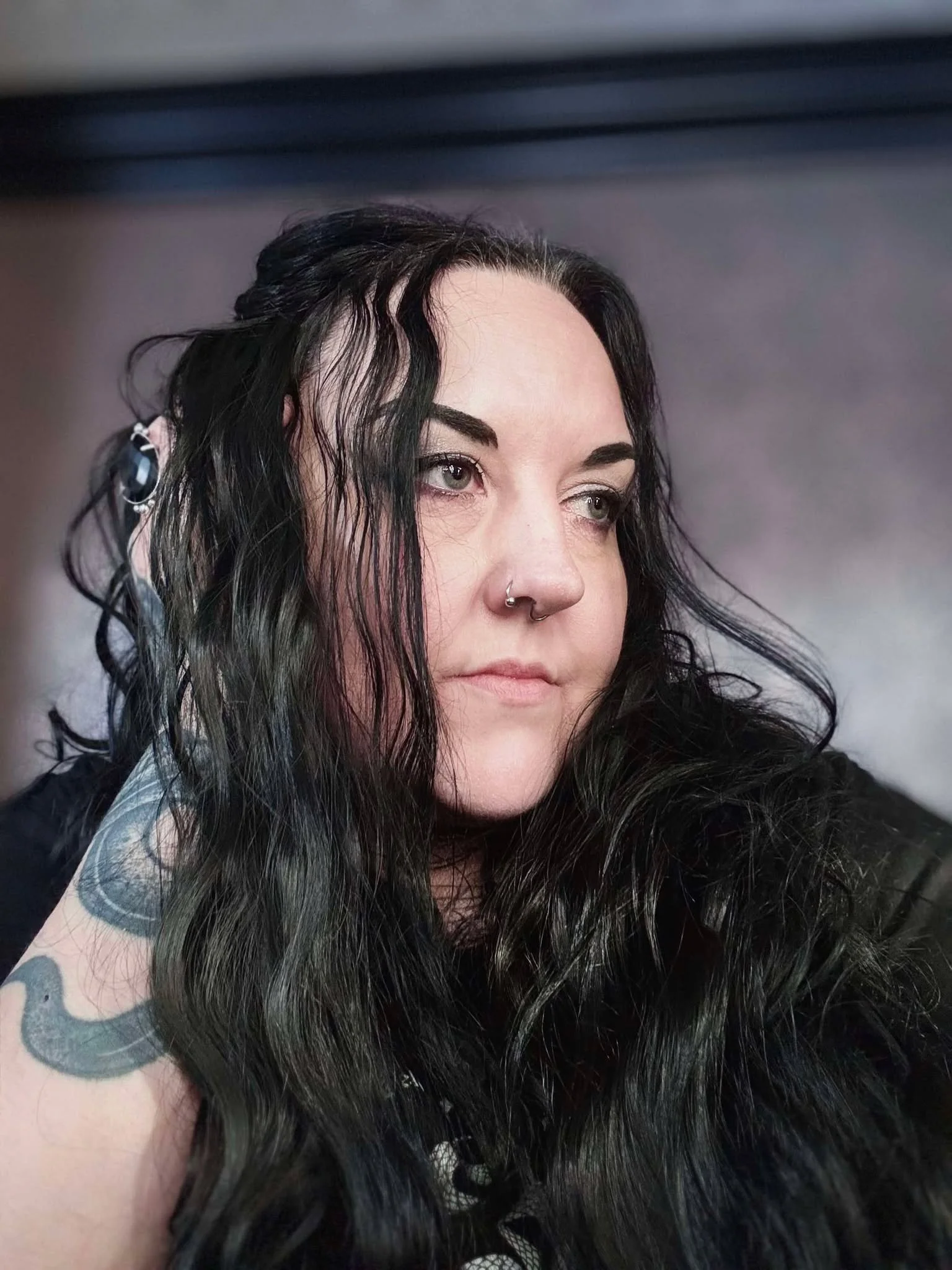 Close-up of a woman with long black wavy hair, a nose piercing, and a tattoo on her arm.