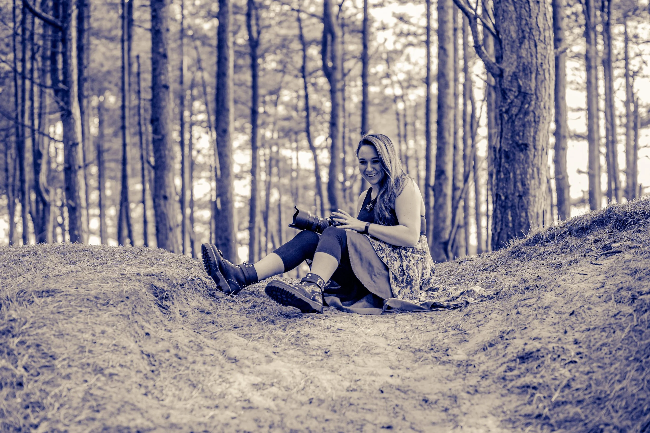 A woman sitting on a dirt path in a forest, holding a camera and smiling.