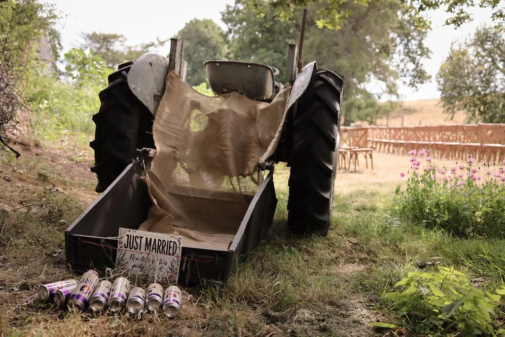 A vintage tractor with a broken wheel in a grassy outdoor setting. There is a 'Just Married' sign in the tractor's tray, along with several cans tied together with string, and pink flowers blooming nearby.