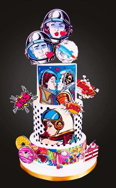 A colorful cake decorated with pop art style images of astronauts, romantic scenes, and comic book elements like speech bubbles and sound effects.