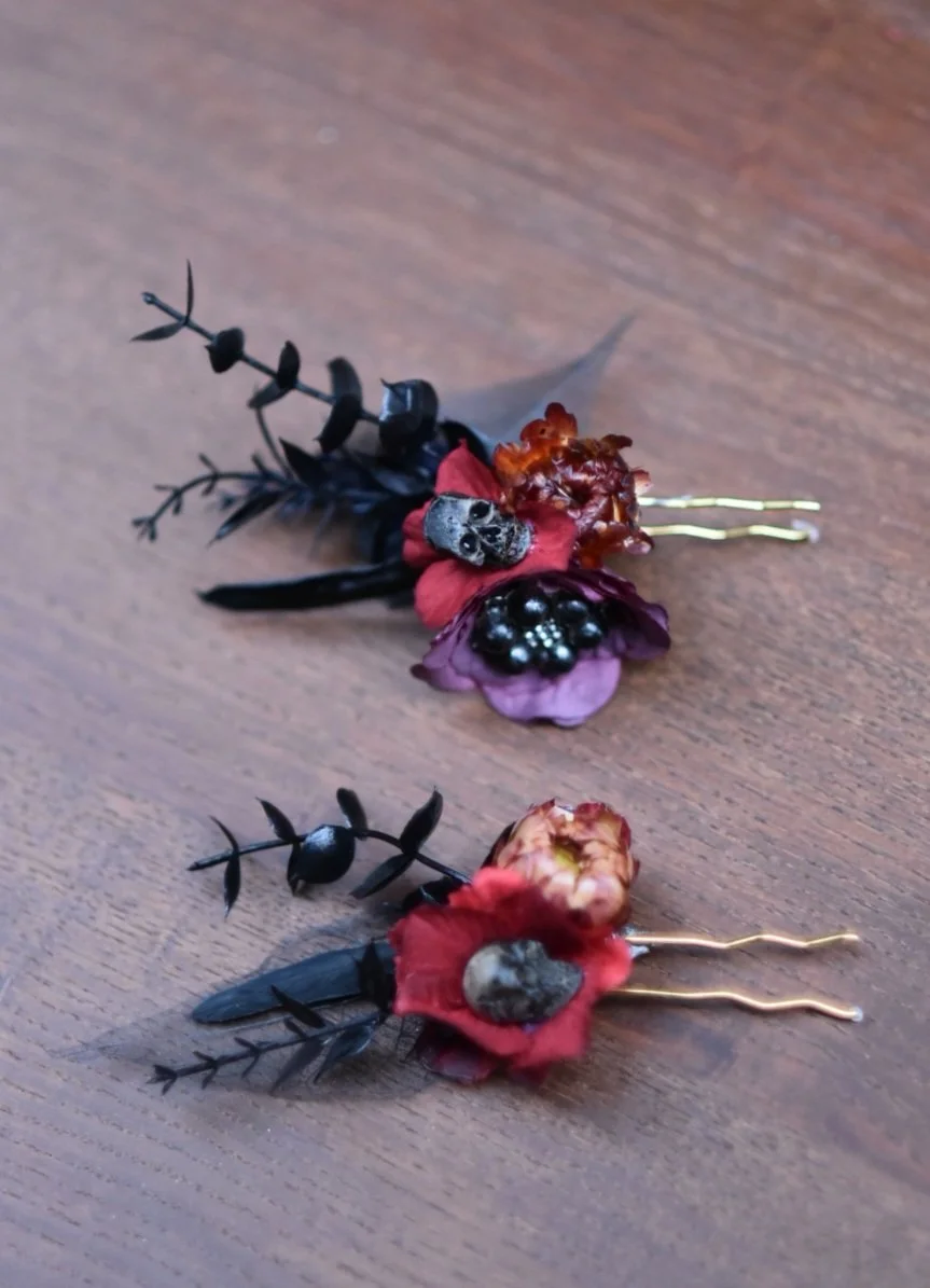 Two decorative brooches made from black leaves, red and purple flowers, a small skull, and black beads, placed on a wooden surface.