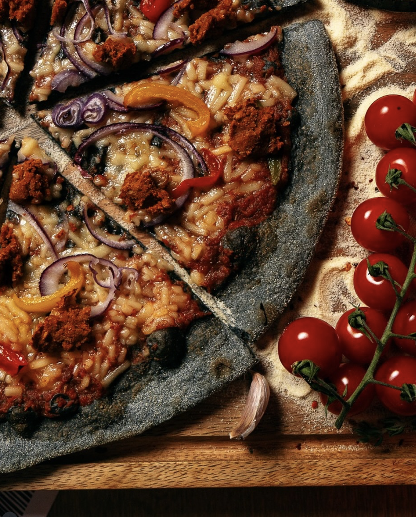 Close-up of a stone pizza with melted cheese, tomato sauce, pepperoni, and sliced red onions, served alongside cherry tomatoes on the vine and a garlic clove on a wooden surface.