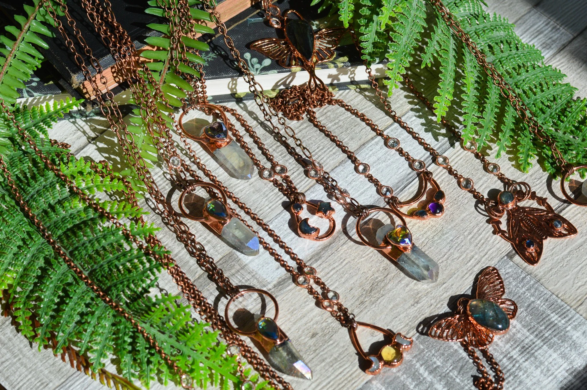 Various copper jewelry pieces including necklaces, pendants, and earrings with gemstones, displayed on wooden surface with green fern leaves.