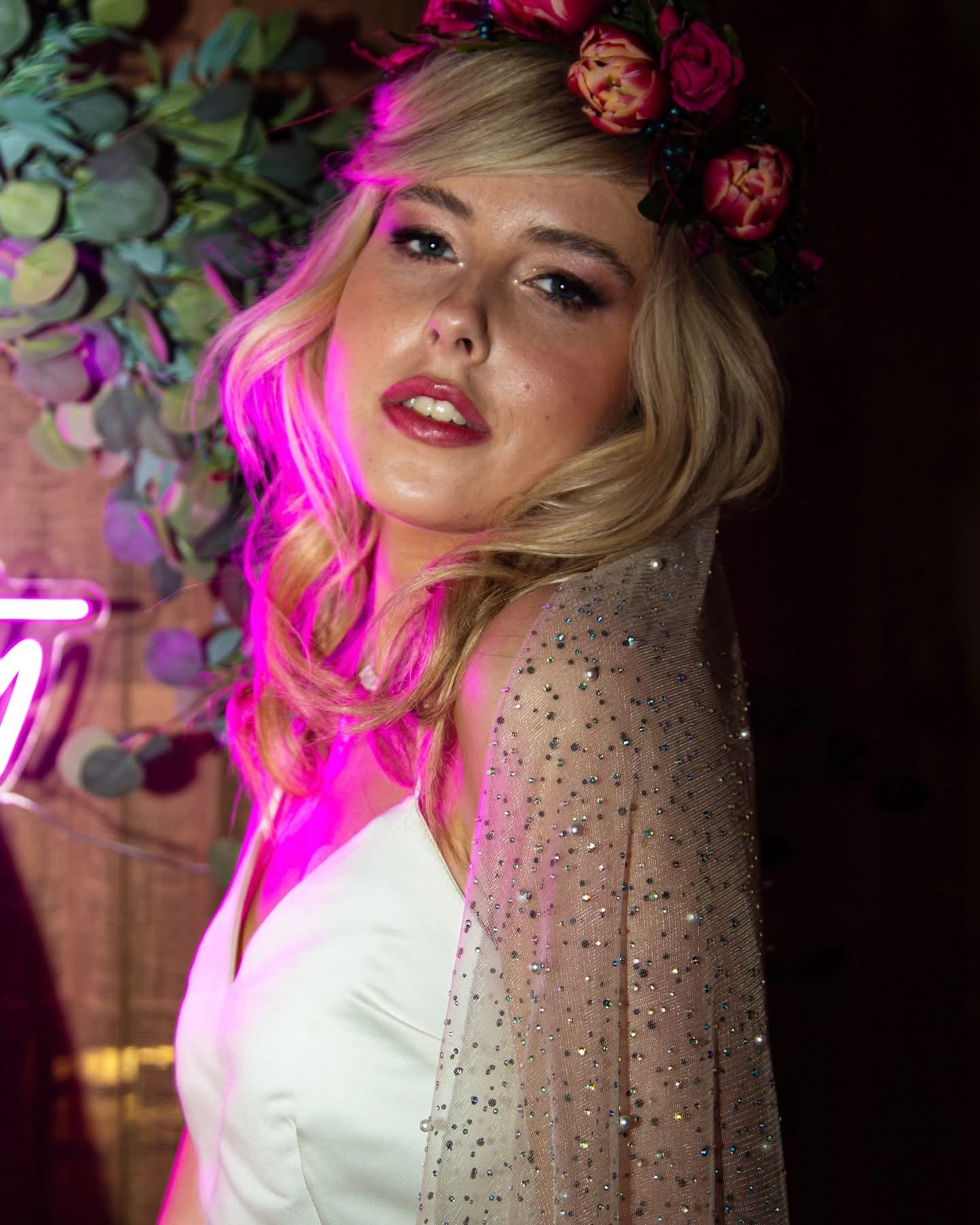 A woman with blonde hair wearing a floral headband, a white dress, and a glittering cape, posing in front of a backdrop with green foliage and pink neon lighting.