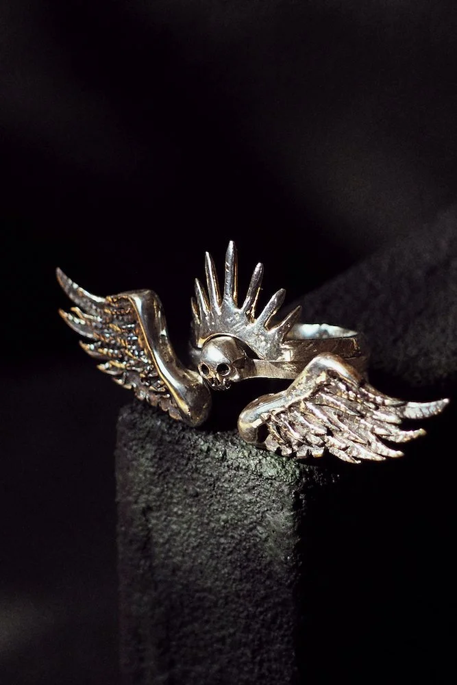 Silver ring with a skull, wings, and spike design, displayed on a black textured surface.