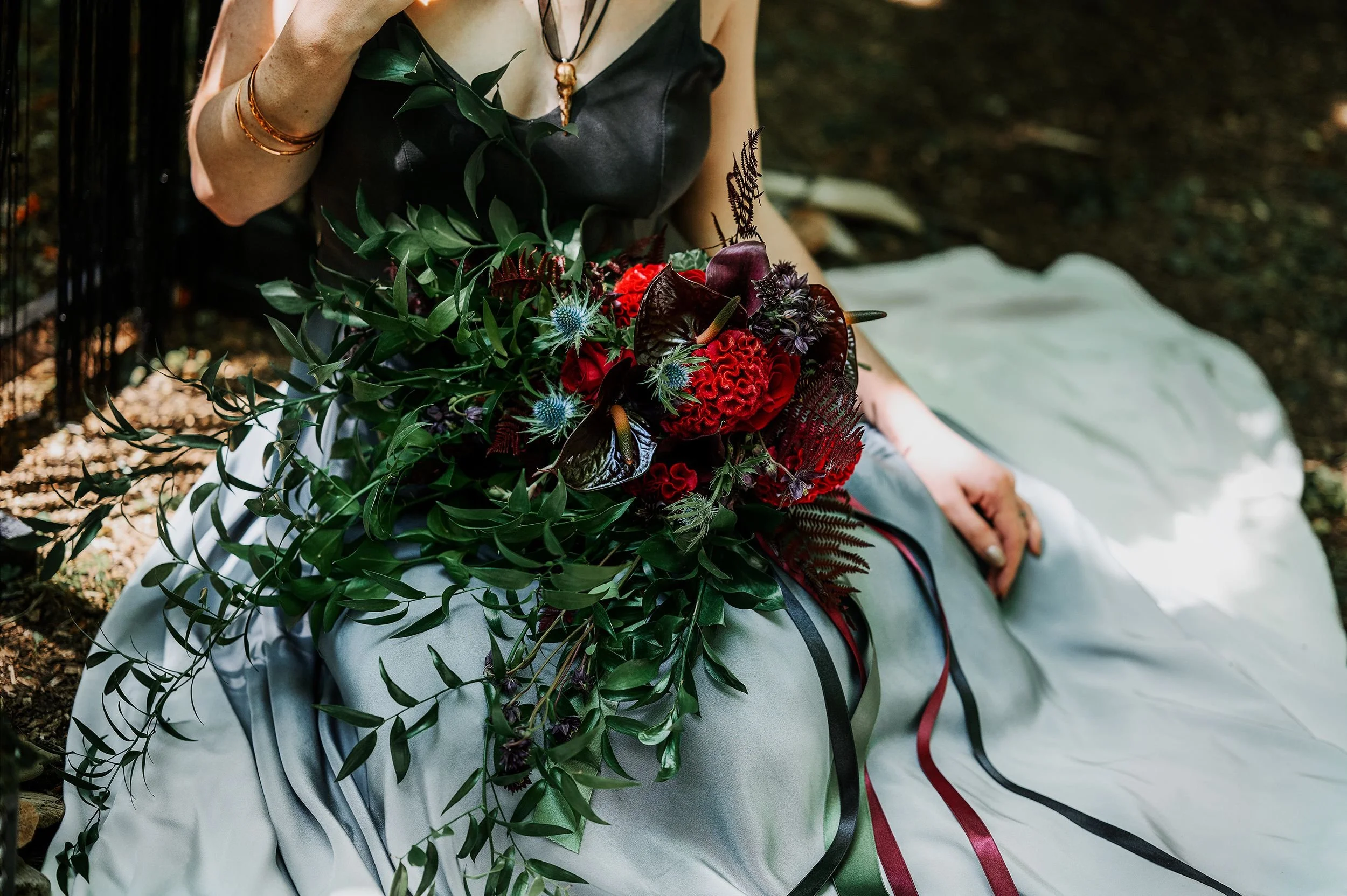 Close-up of a woman sitting outdoors on a white cloth, holding a large bouquet of dark red, black, and purple flowers with green foliage. She is wearing a black top, gold bracelets, and a necklace with a pendant.