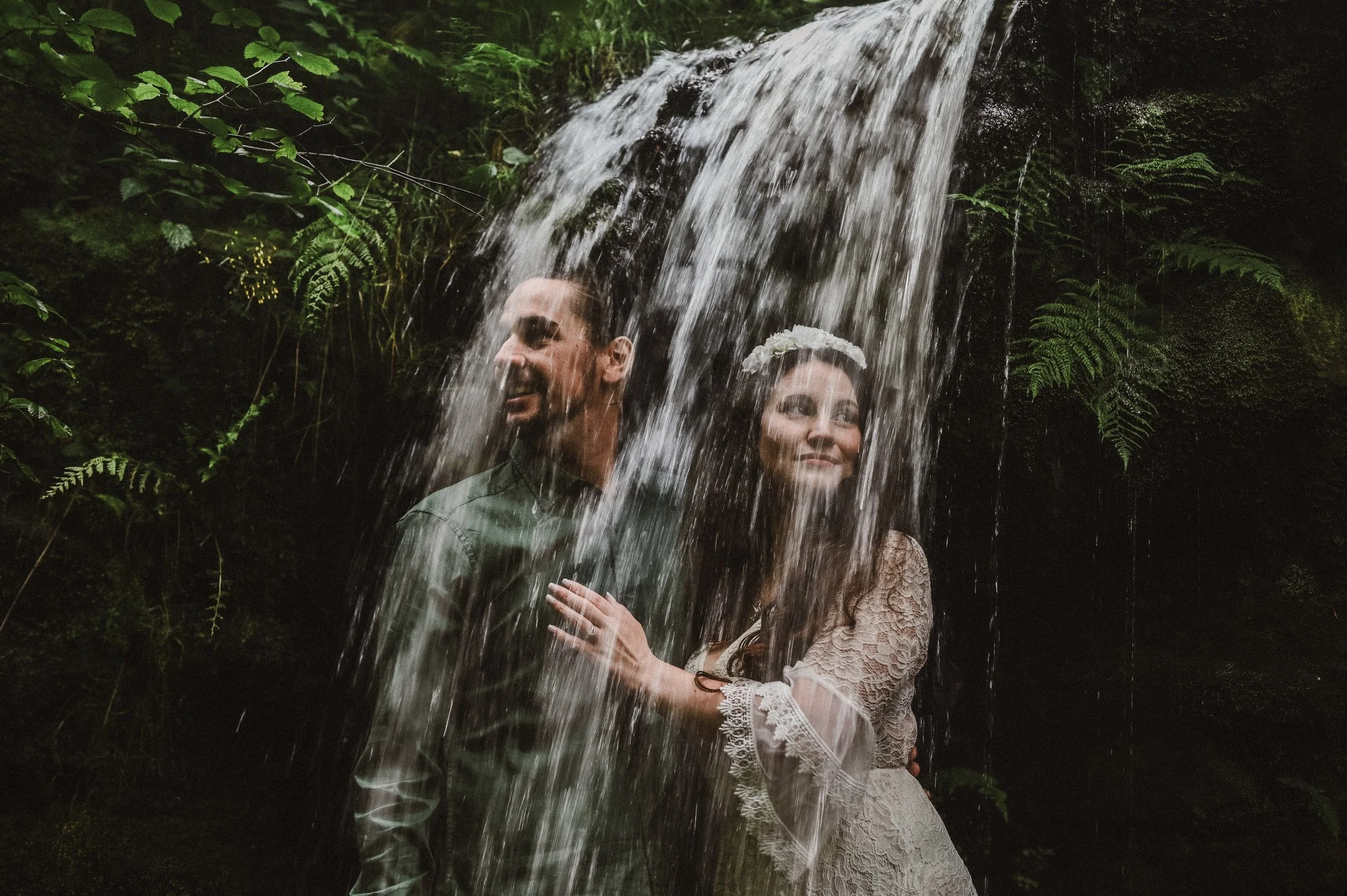 A couple standing under a waterfall in a forest setting, smiling and enjoying a moment of intimacy.