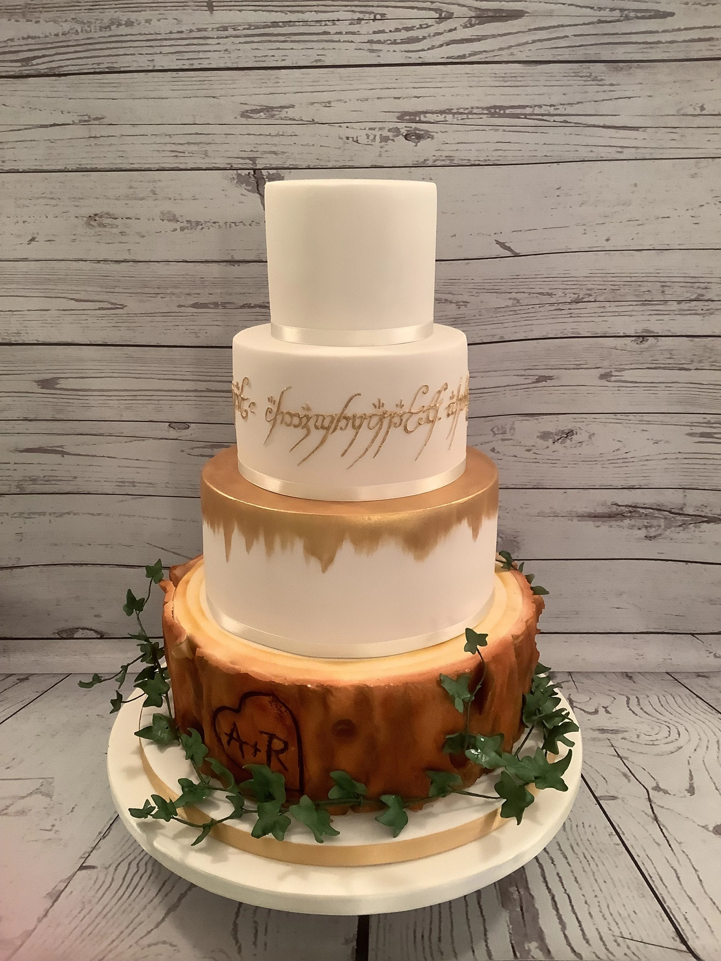 Three-tiered wedding cake with a wood log base, decorated with green leaves, featuring gold accents, California: A+R inside a heart on the log base, and gold piping and lettering on the upper tiers, set against a wooden backdrop.