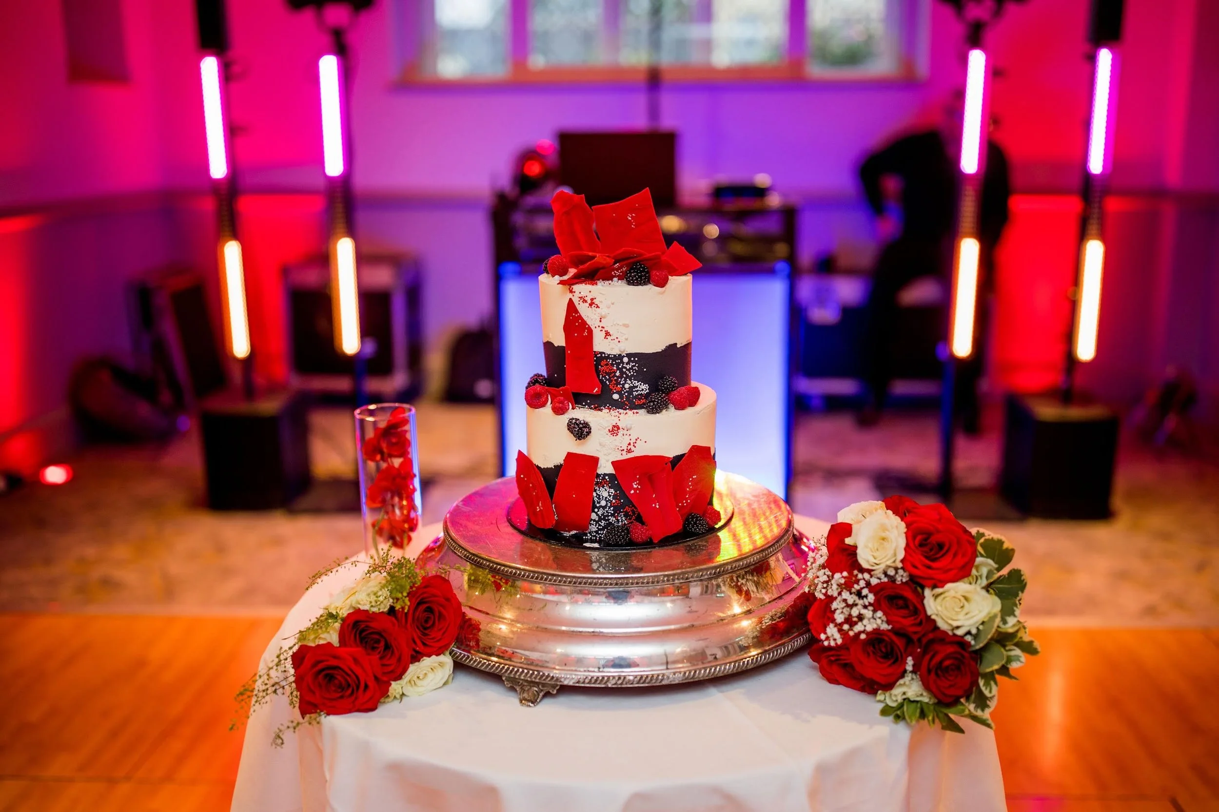 A two-tiered wedding cake decorated with red and black accents, roses, and red chocolate shards, placed on a silver cake stand with floral arrangements of red and white roses on each side. The background features colorful stage lighting and a DJ setu