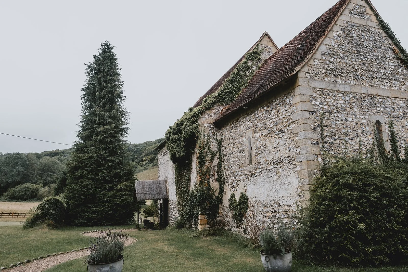 An old stone church with ivy growing on its walls, surrounded by a garden with potted plants, bushes, a tall evergreen tree, and a gravel walking path, under a cloudy sky.