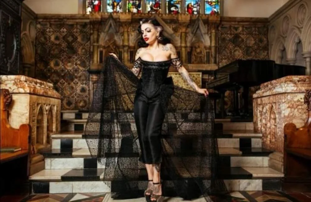 A woman dressed in black lace and satin sitting-hiking dress, standing on a marble staircase inside a church or cathedral with ornate wooden and stone architectural details, stained glass windows, and a grand piano in the background.