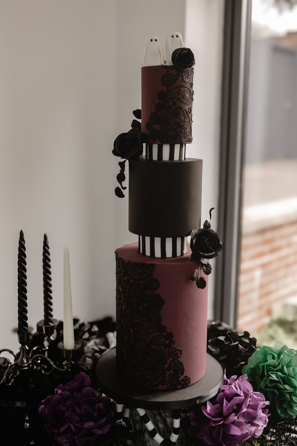 A four-tiered cake decorated with black lace, black roses, and black and pink accents, positioned near a window.