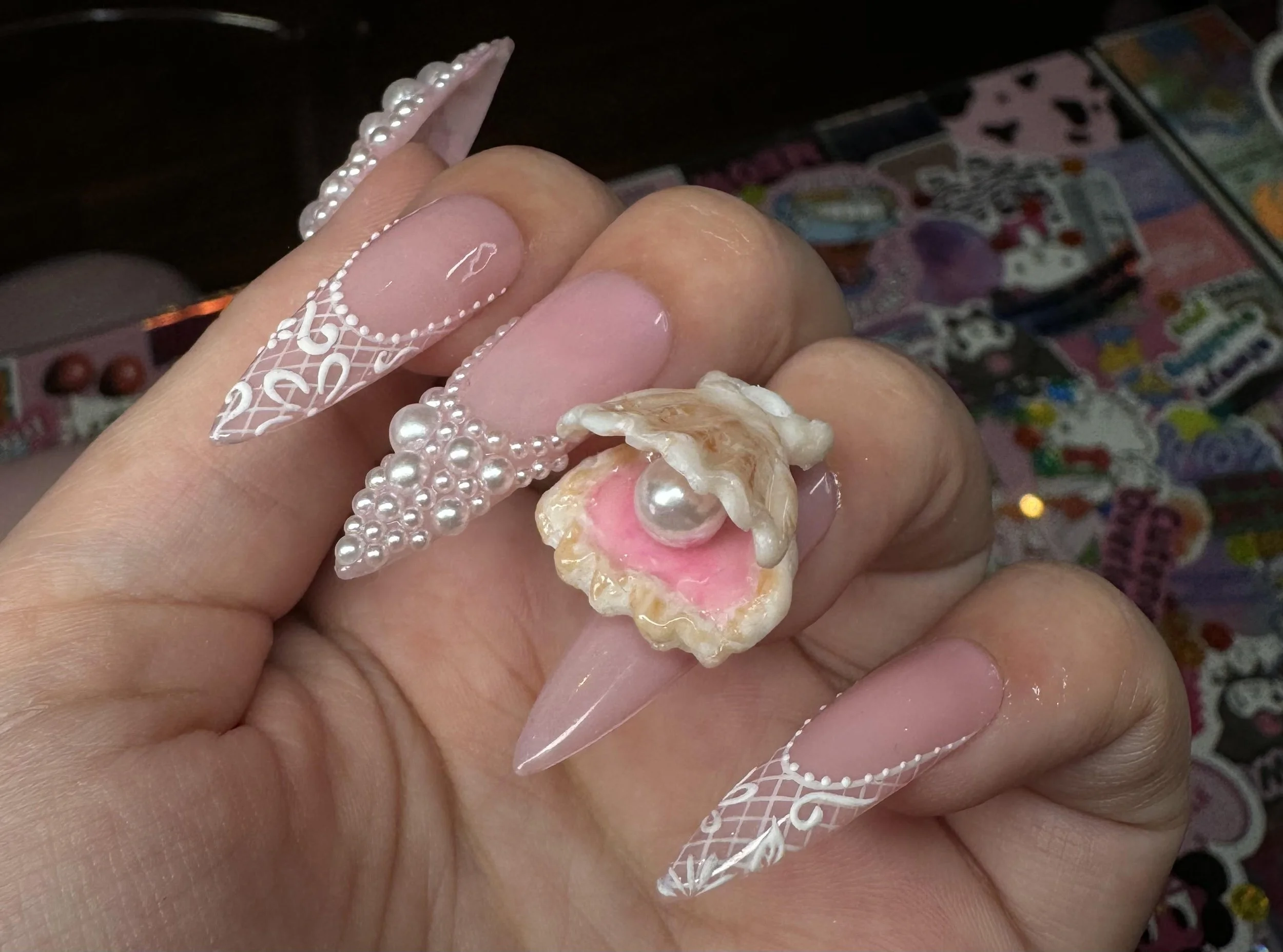 Close-up of a hand with long, decorative, pink and white nails, holding a pearl ring and a shell-shaped ring with a pearl inside, against a background of colorful stickers or cards.
